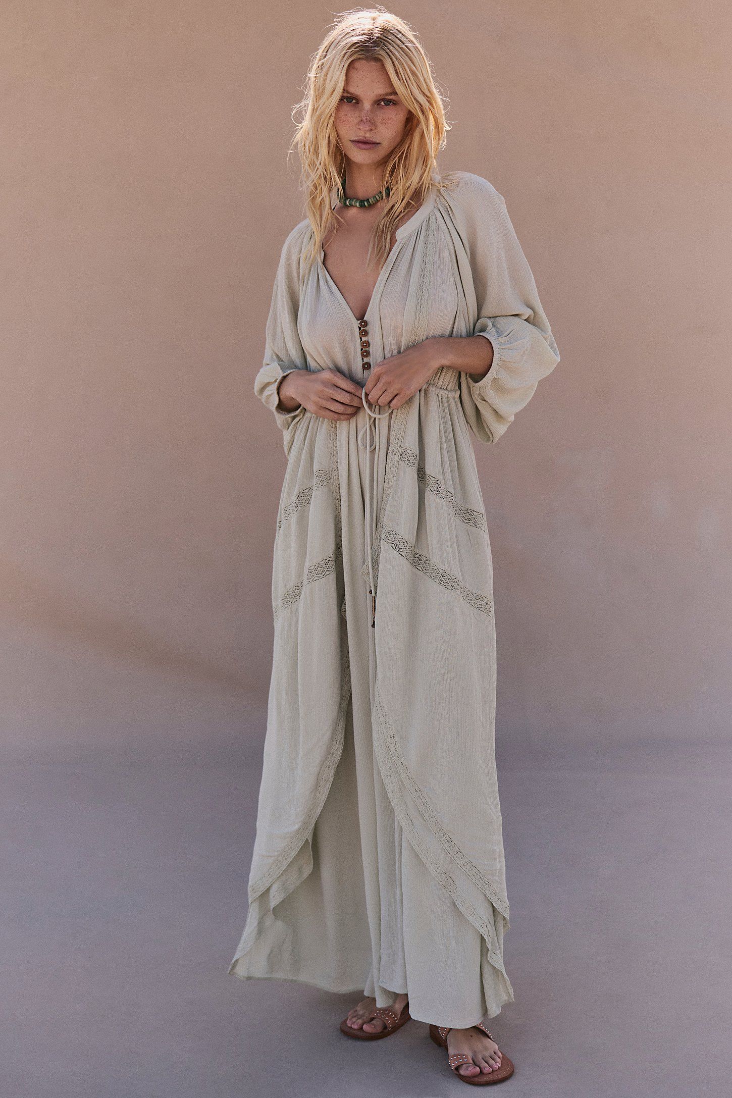 A blonde woman models a flowing beige boho maxi dress with lace trim details and three-quarter sleeves.