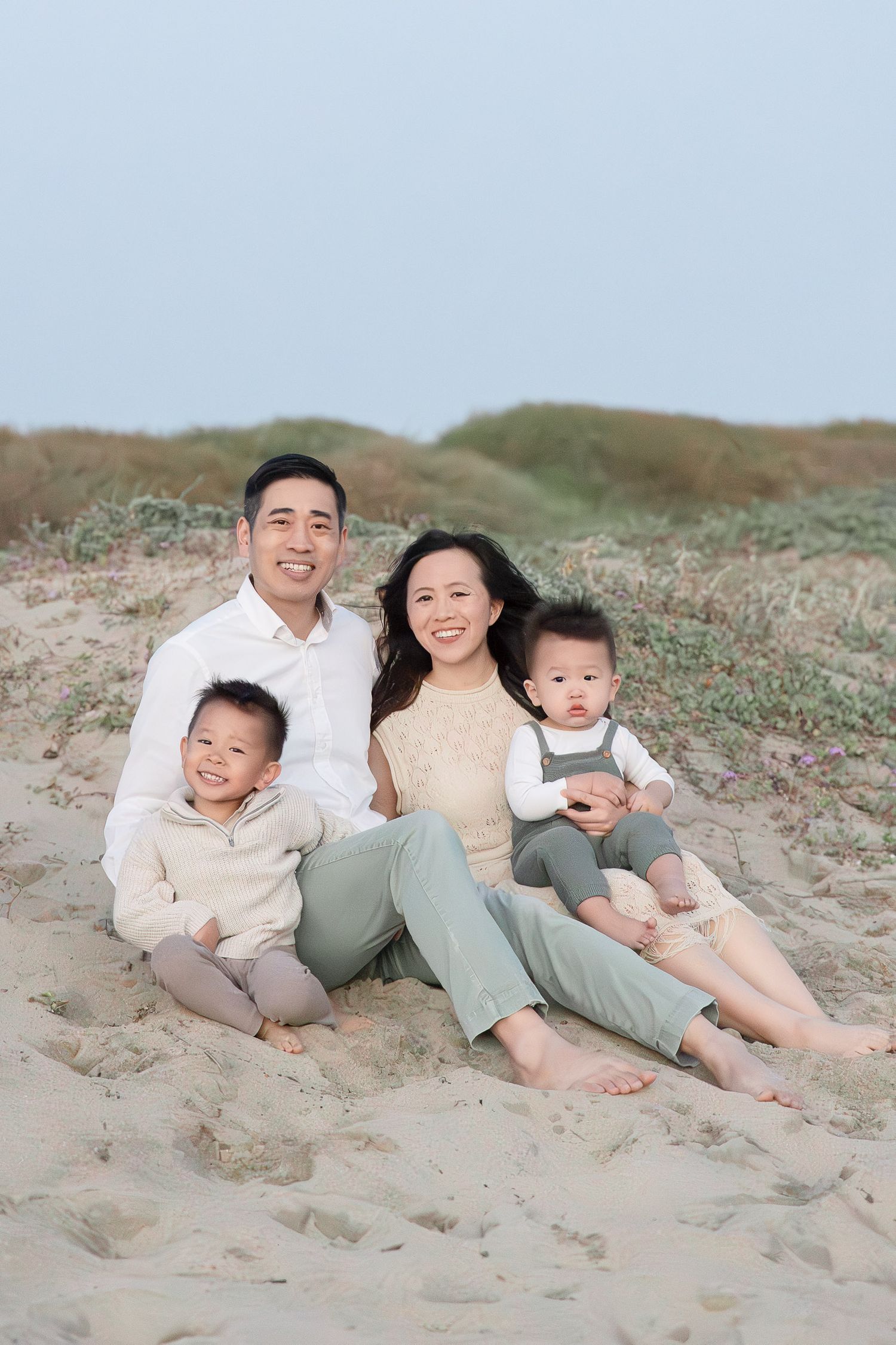 A happy Asian family of four sits together on a sandy beach, smiling warmly for a portrait photo.