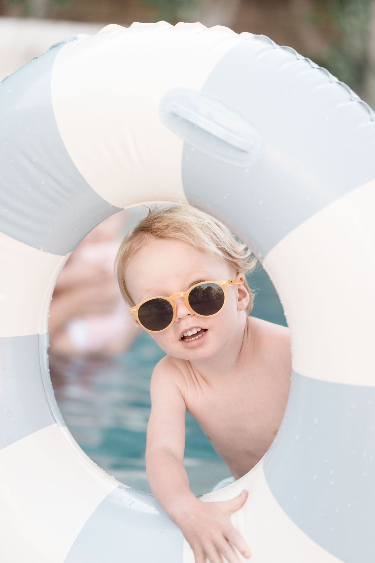 A happy toddler wearing sunglasses peeks through a striped inflatable pool ring in a swimming pool.