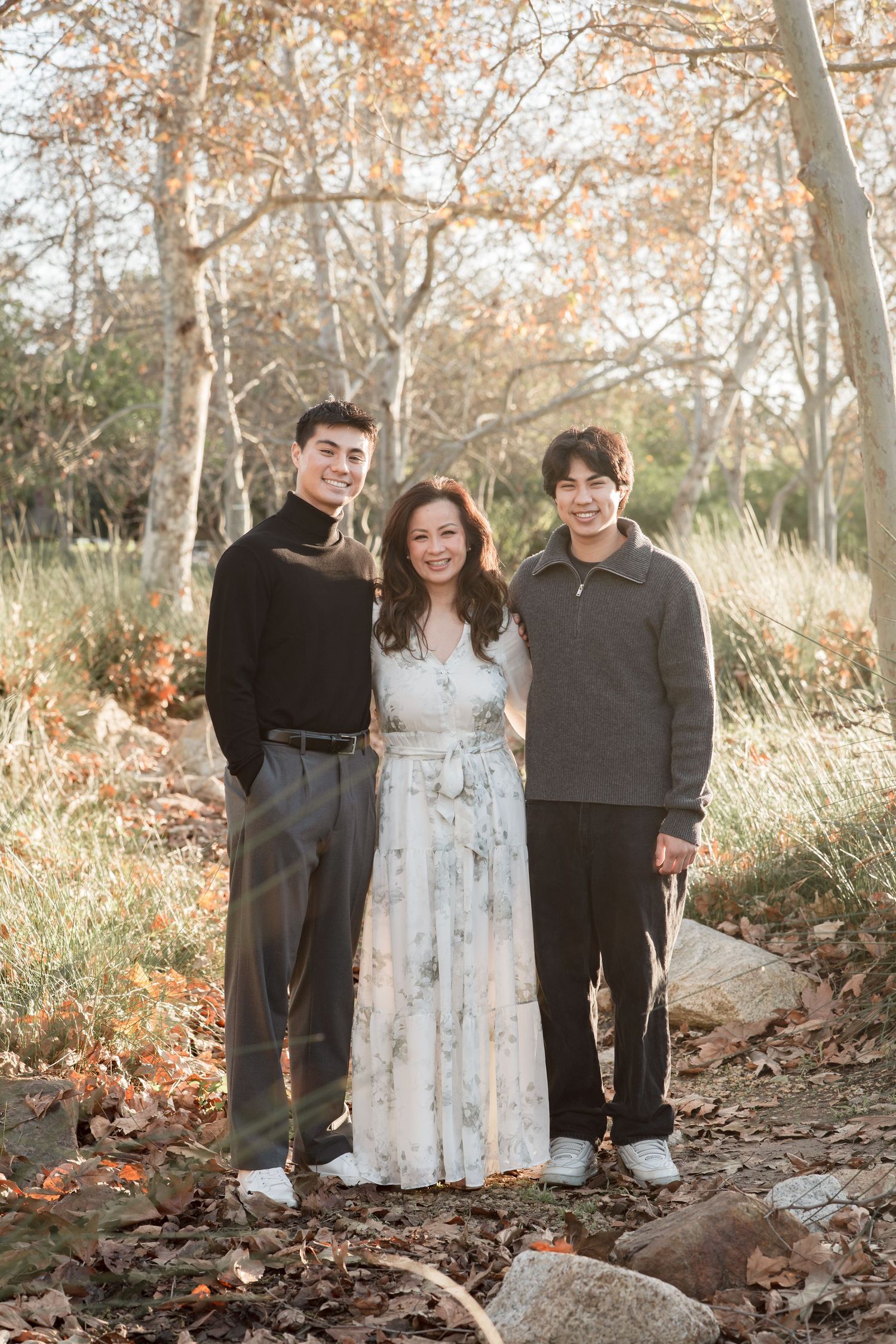 A woman in a floral dress poses with two teenage boys in a sunlit autumn park with fall foliage.