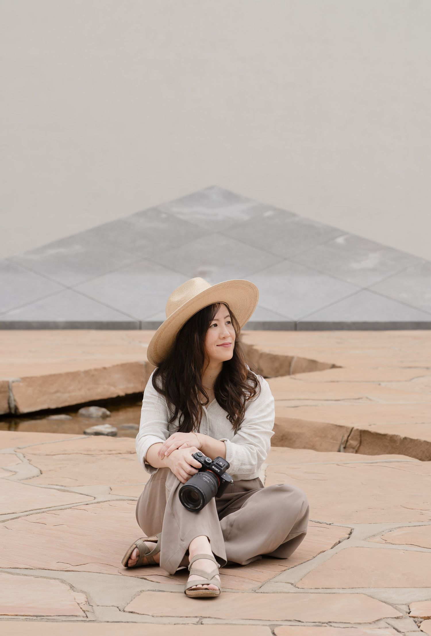 Woman in straw hat sitting cross-legged on stone patio holding camera, smiling outdoors near geometric structure.