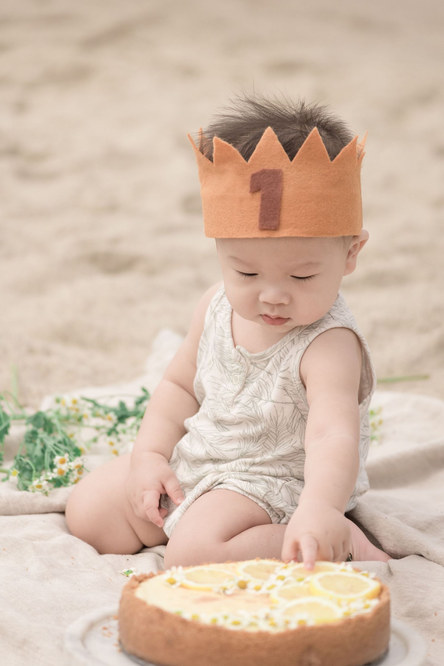 Baby wearing a number one crown celebrating first birthday with a lemon tart on a beach blanket.