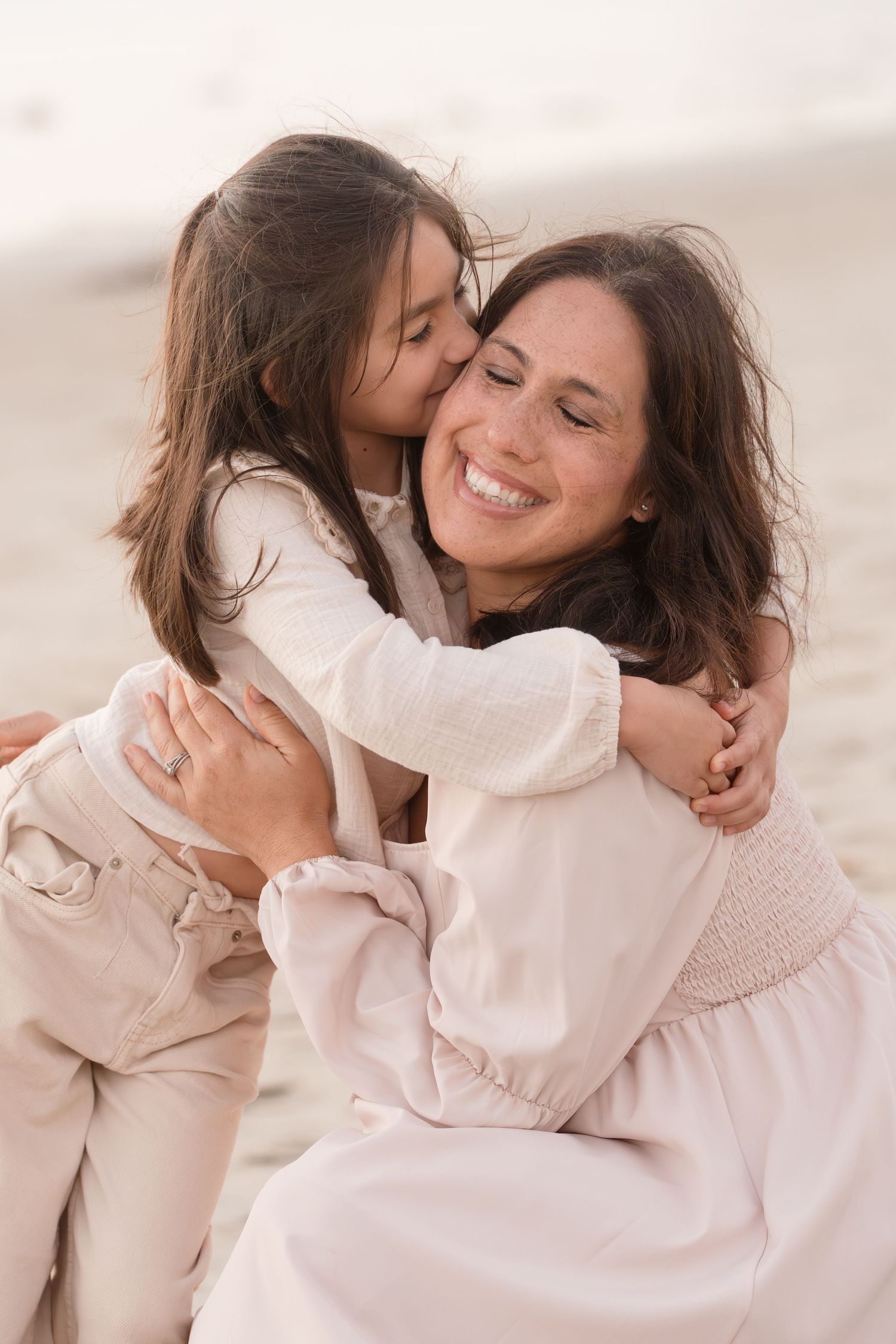 Young girl hugs and kisses her smiling mother on a bright sandy beach.