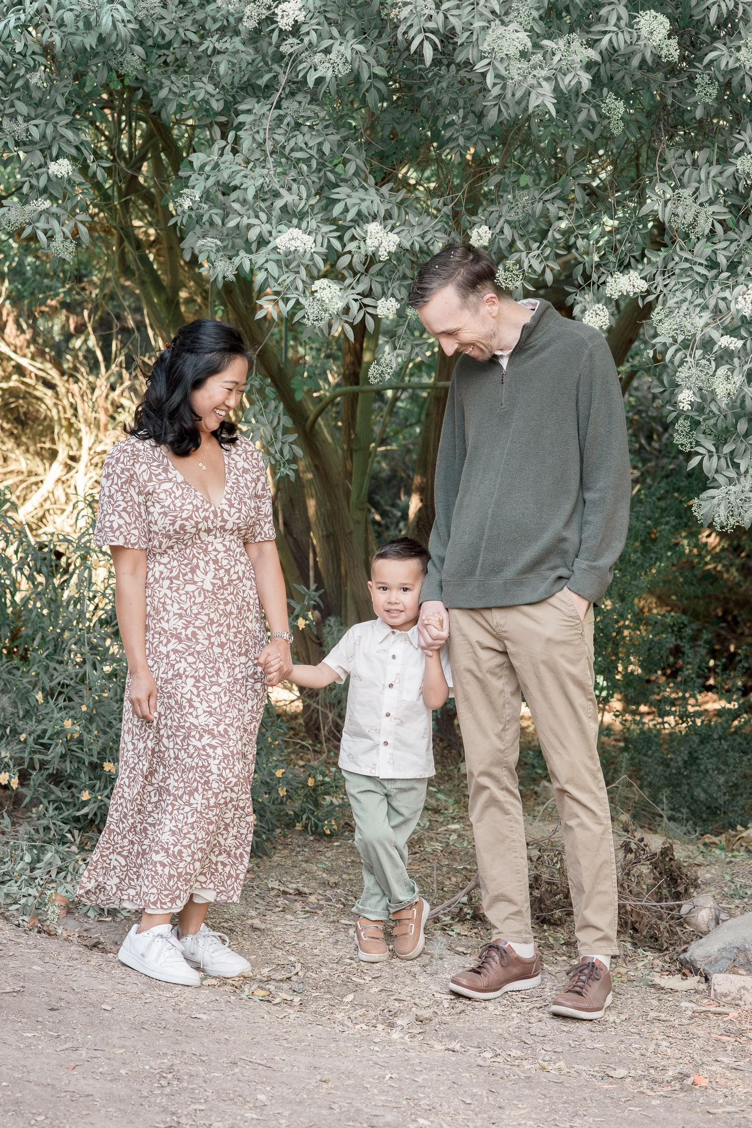 A family of three walks hand-in-hand outdoors, with parents smiling at their young toddler son among lush green trees.