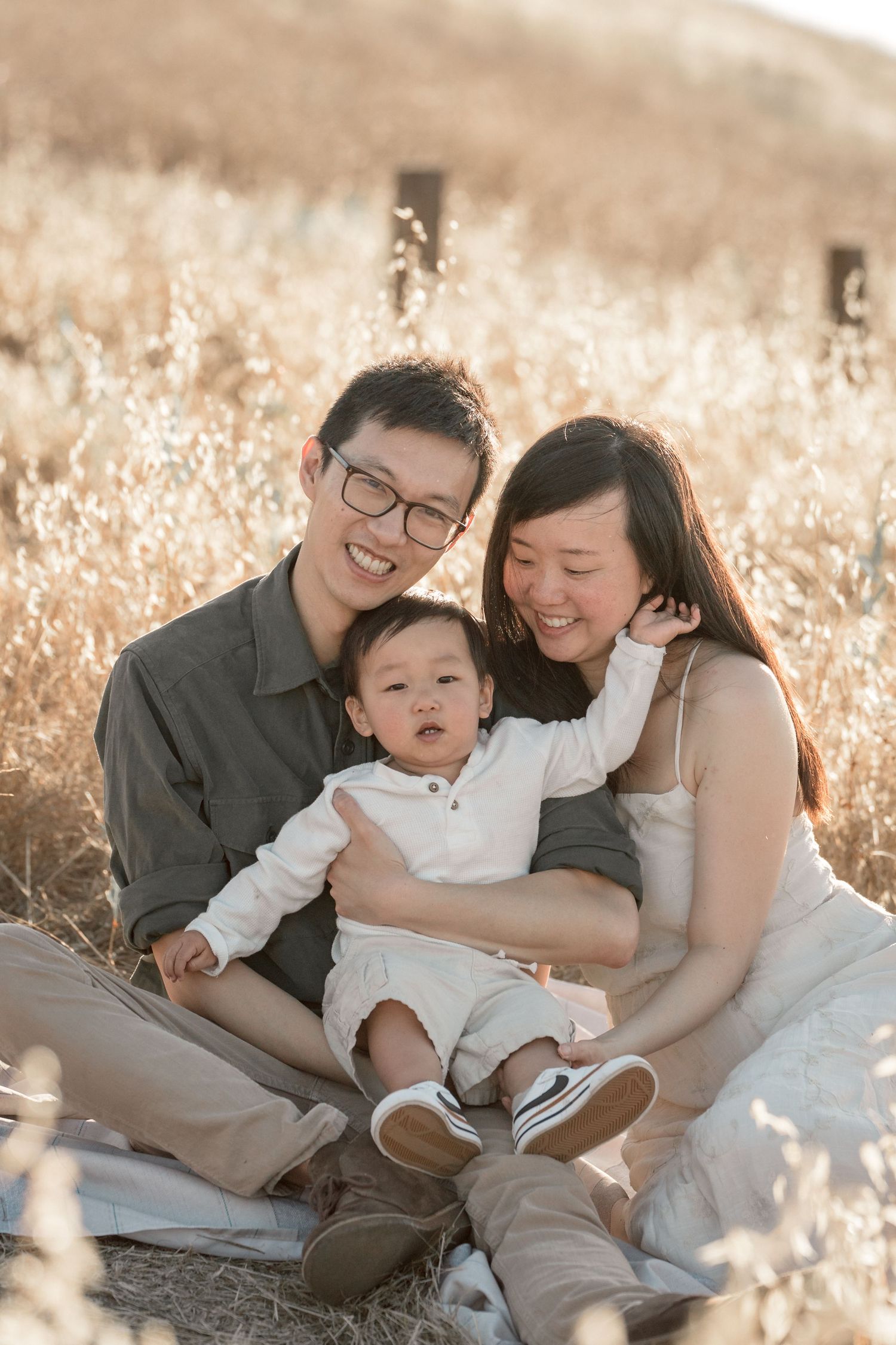 Asian family with parents and baby sitting in golden field, smiling warmly during outdoor portrait session.