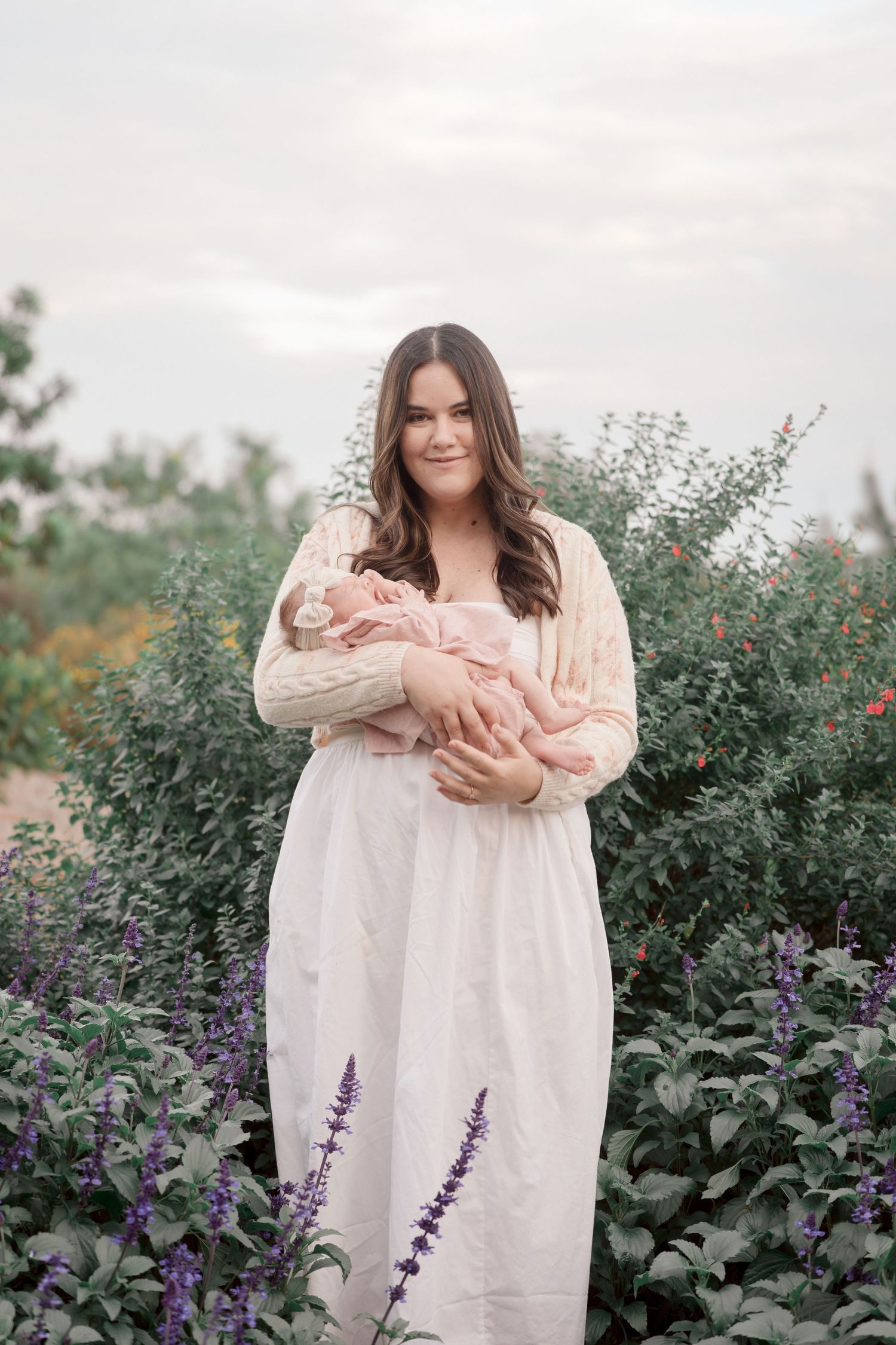Mother in white dress and lace cardigan holding newborn in pink bow, standing among purple lavender and green bushes.