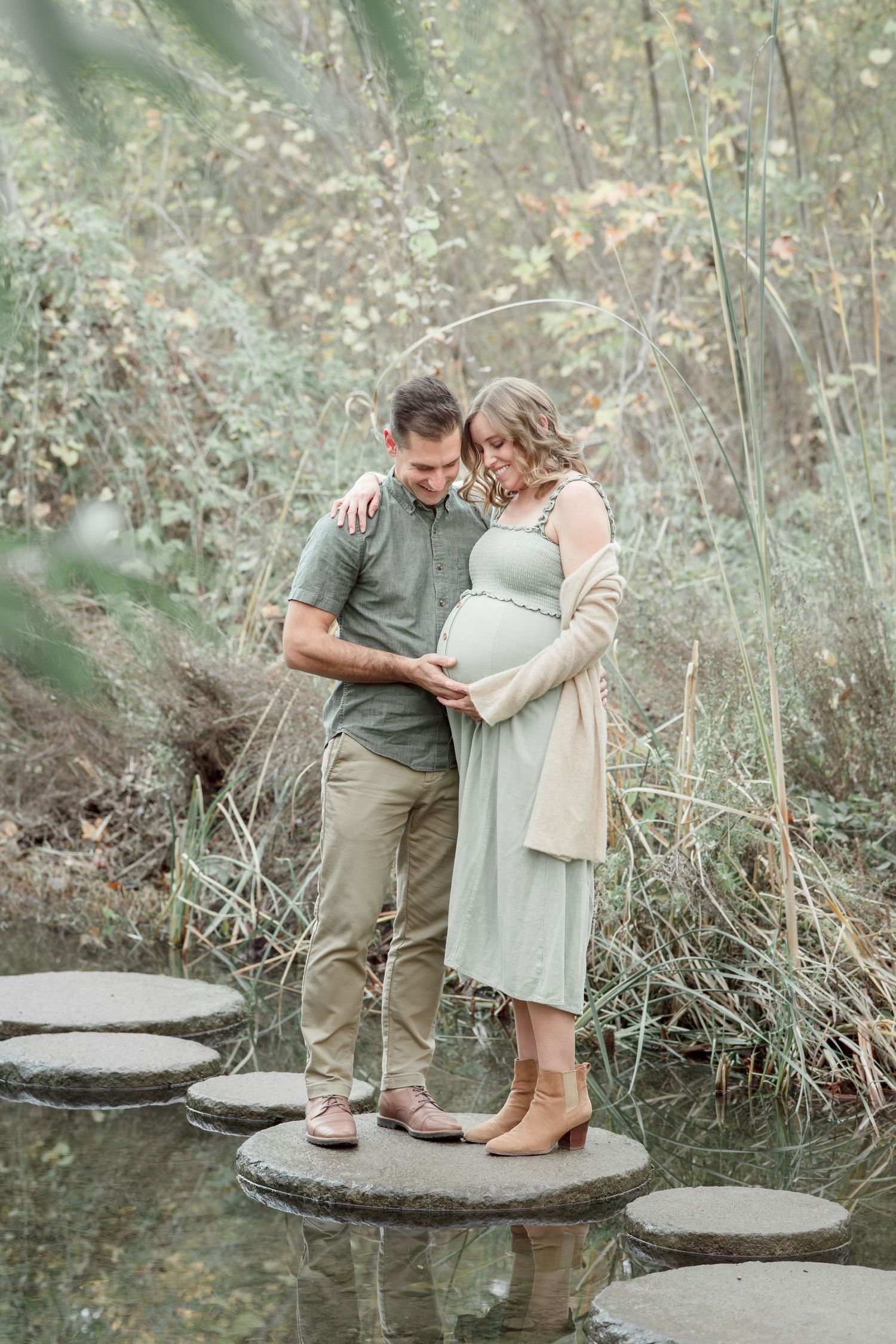 A couple stands on stepping stones over a pond, lovingly cradling a pregnant belly in nature.