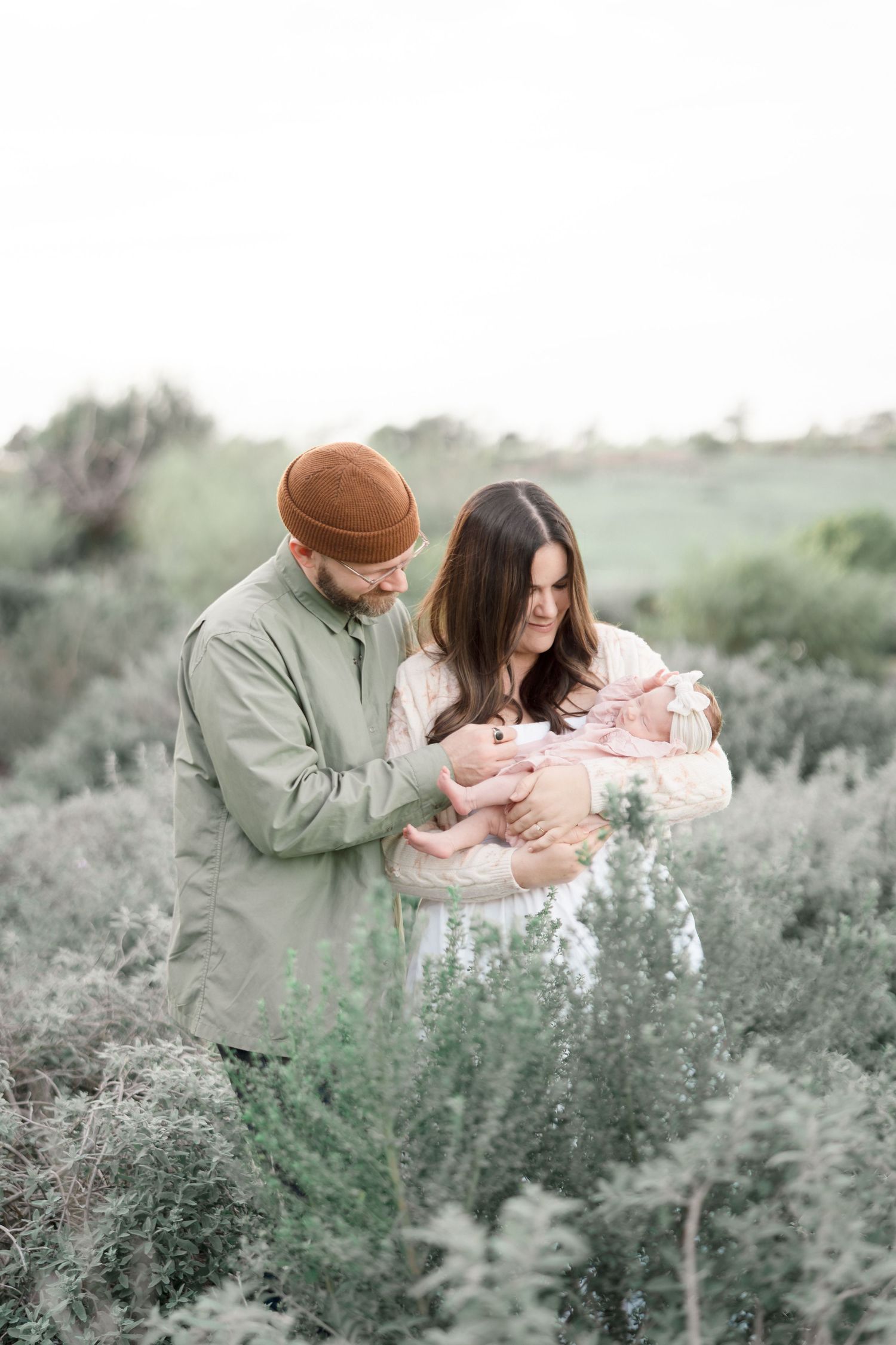 New parents in a green field tenderly gaze at their newborn baby wrapped in a pink blanket.