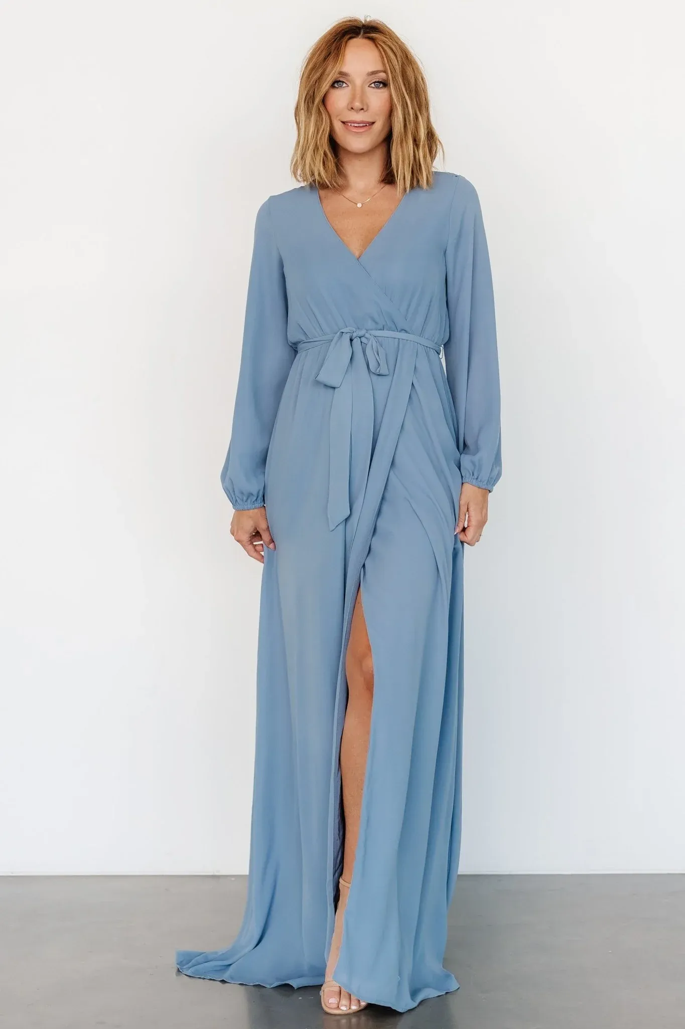 Woman wearing a dusty blue long sleeve wrap maxi dress with a front slit and waist tie.