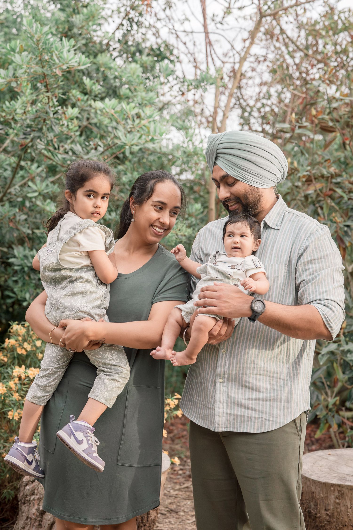 A Sikh family of four poses outdoors, parents holding a toddler and baby amid lush green garden foliage.