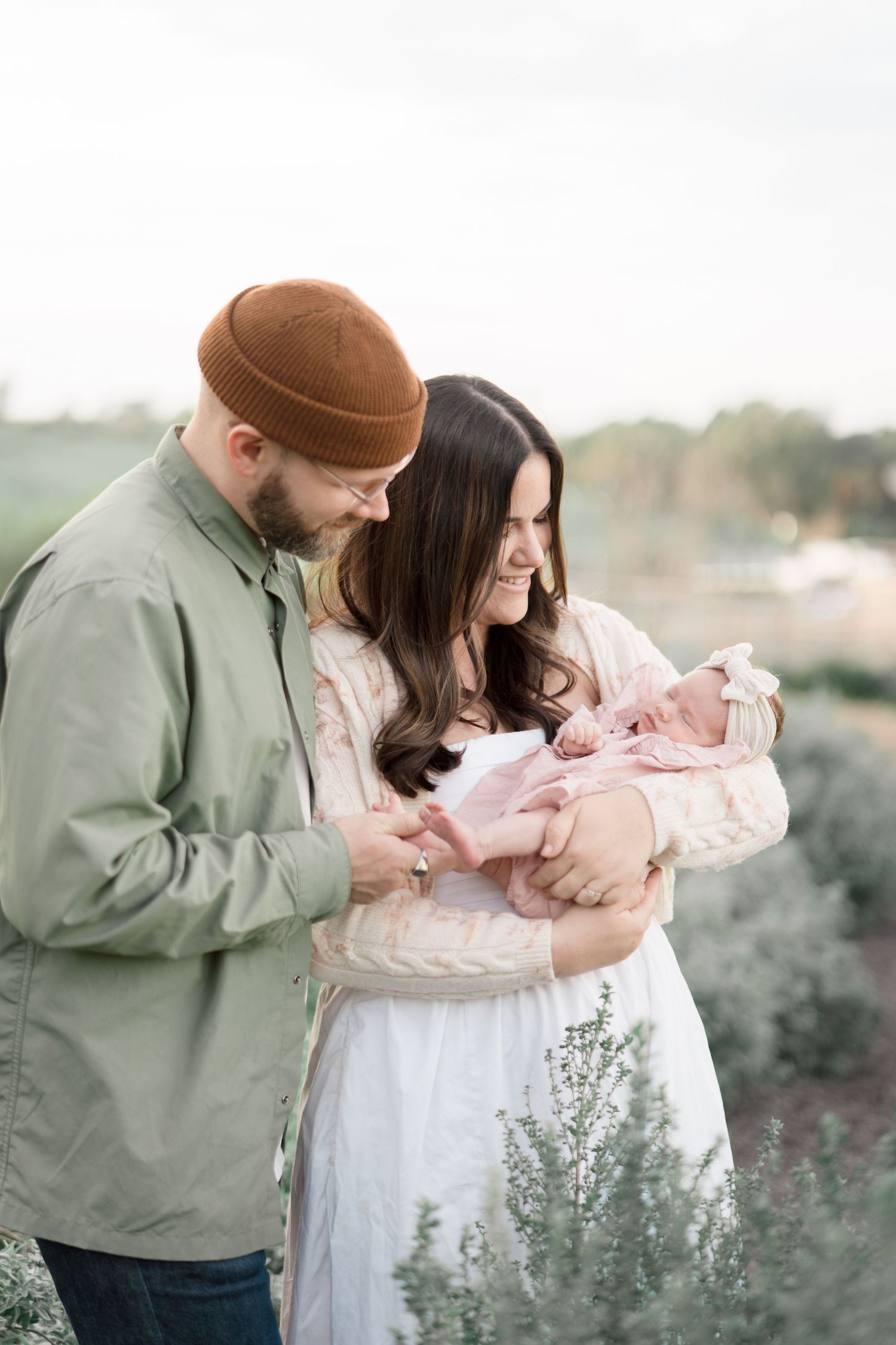 New parents in outdoor field smiling down at newborn baby dressed in pink.