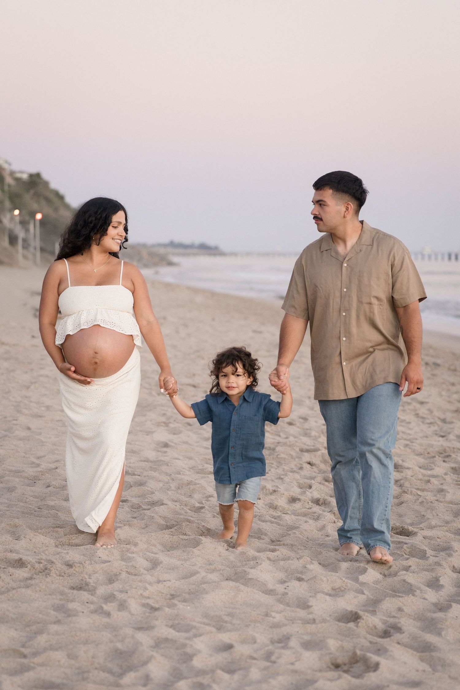 A pregnant woman and her partner walk barefoot on the beach holding their young son\u2019s hands.