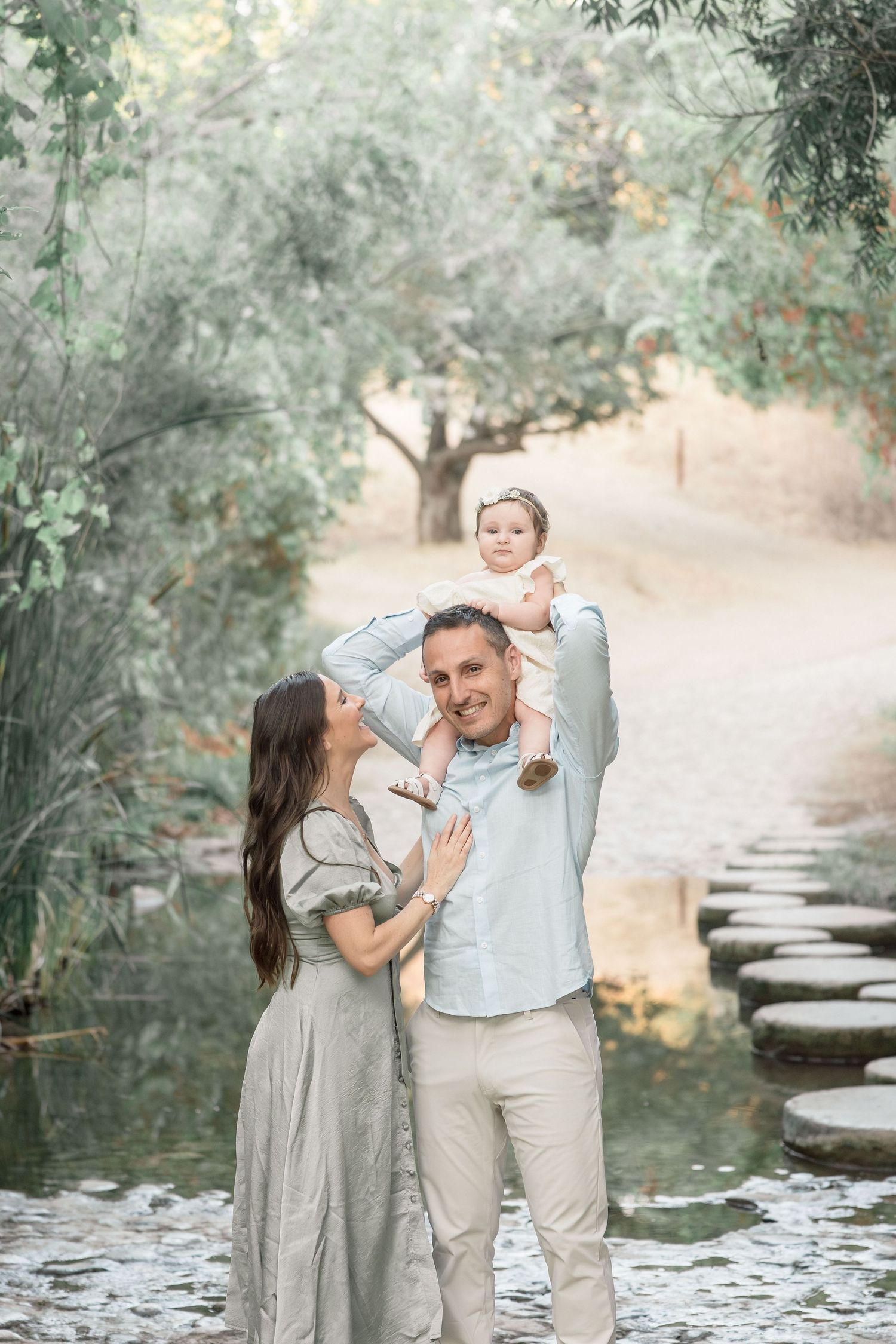 Happy family with father lifting baby overhead while mother smiles beside stepping stone path by pond.