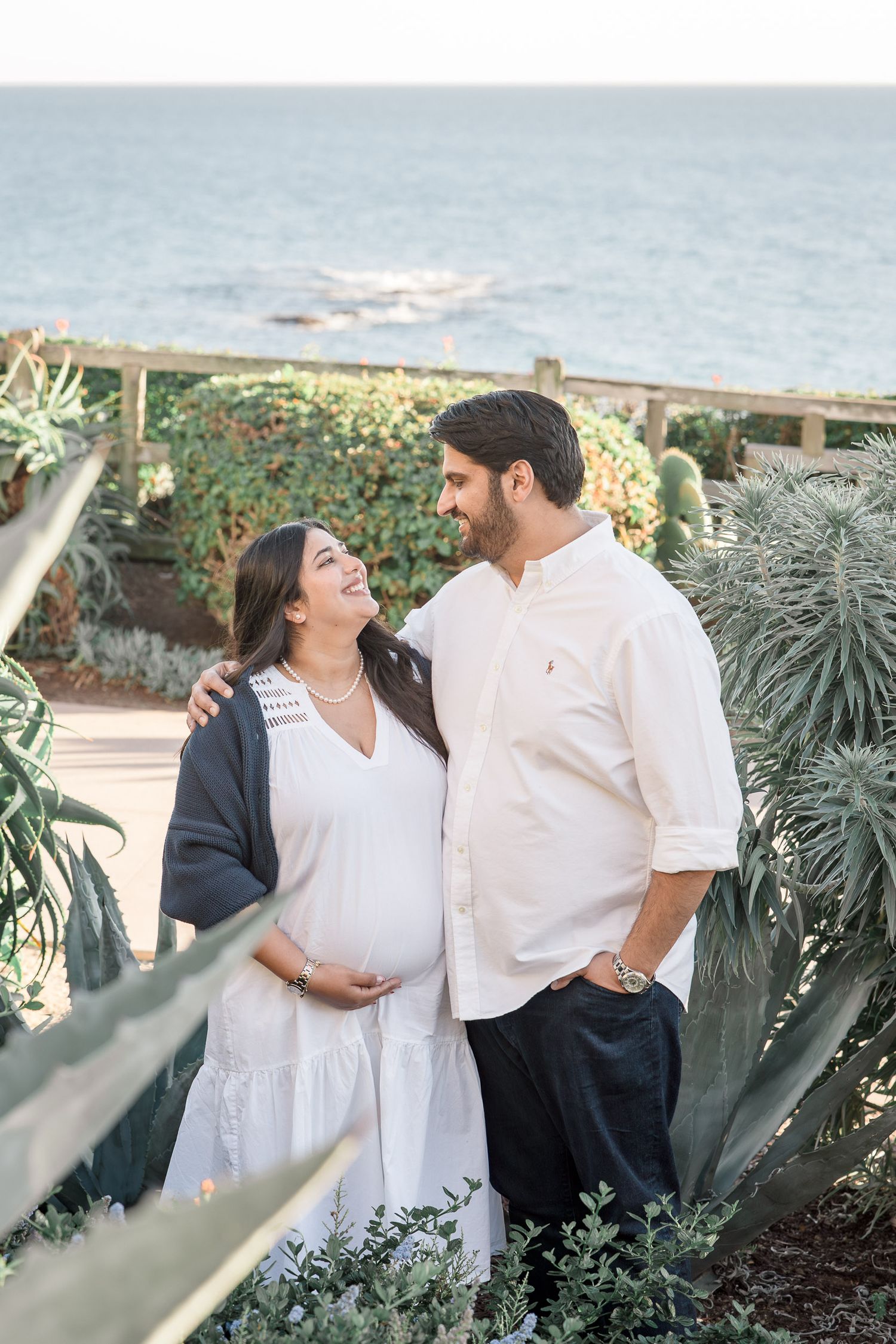 Pregnant couple smiling at each other in a coastal garden, woman in white dress holding baby bump, man in white shirt.