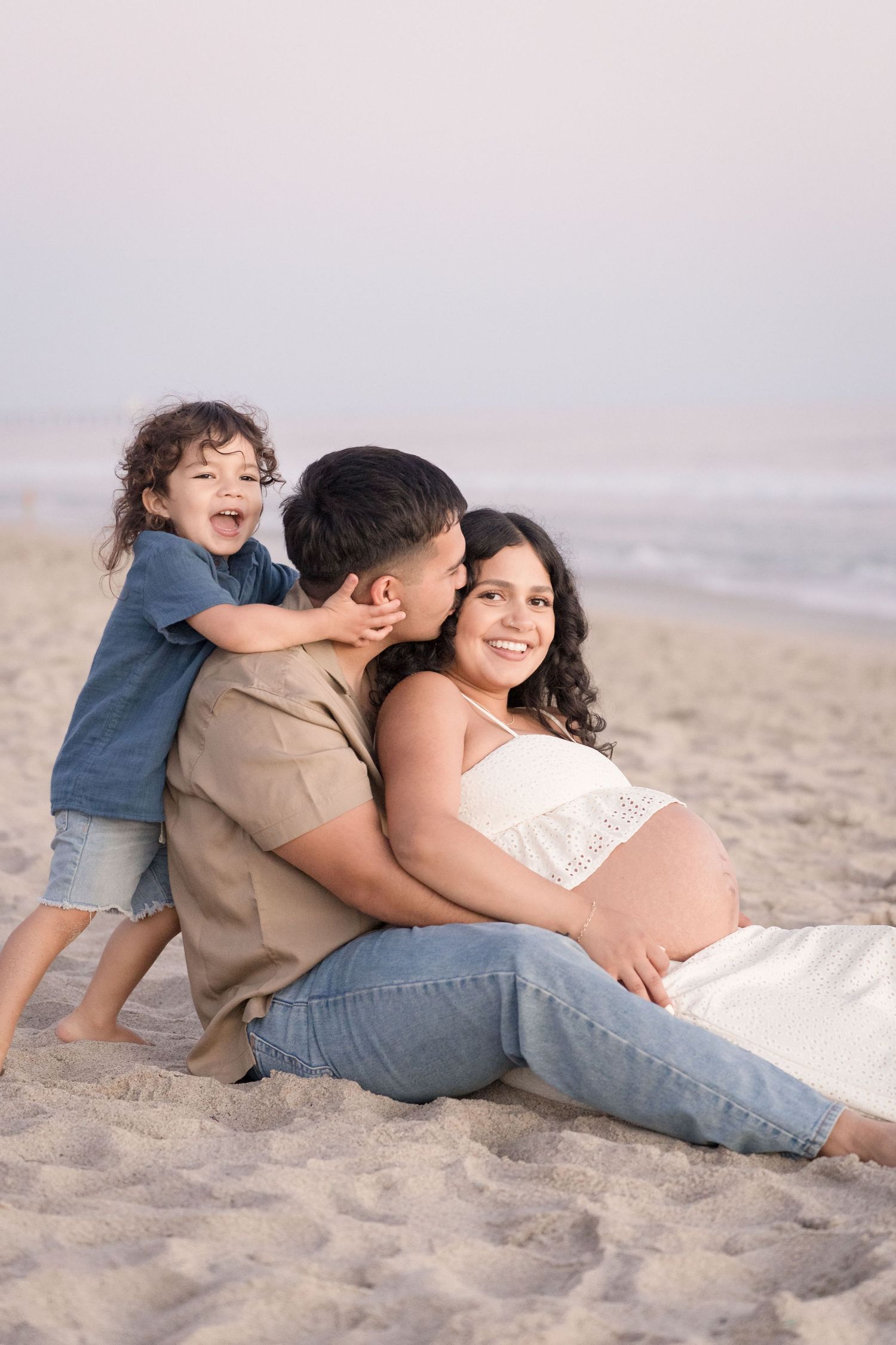 A happy pregnant family sits together on the beach at sunset, with their laughing toddler son.