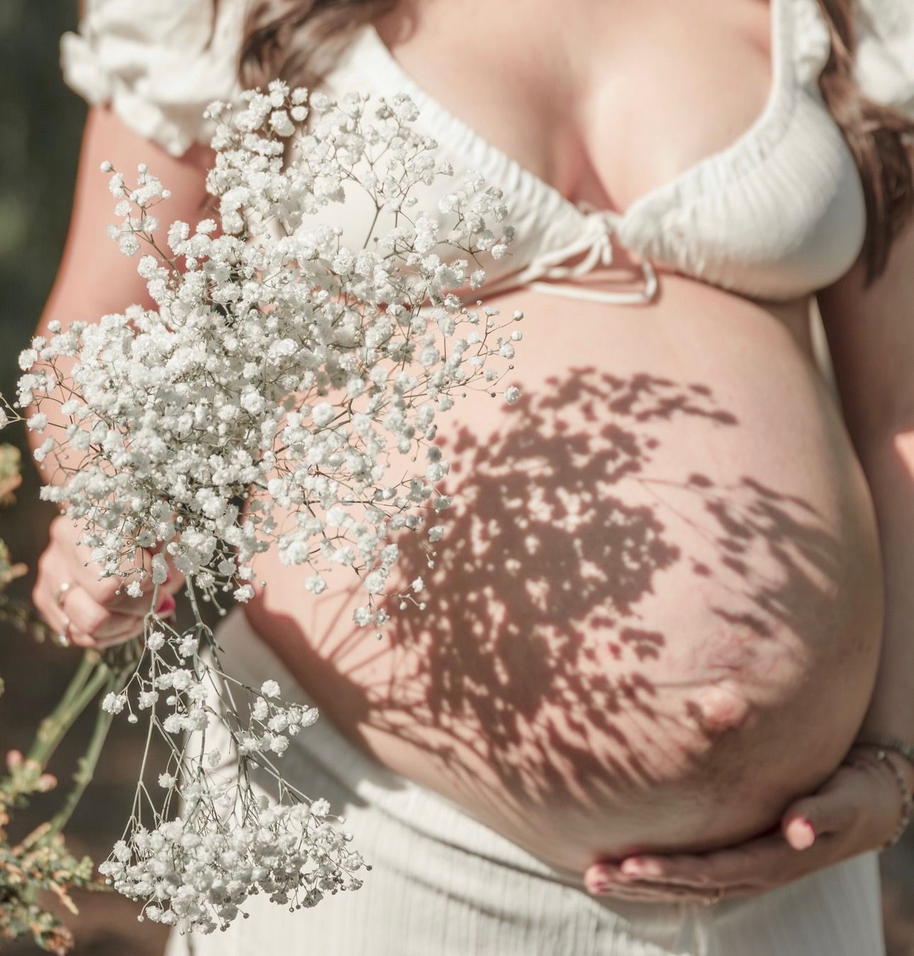 Pregnant woman in white dress holding delicate baby's breath flowers with sunlight casting floral shadows on her bare belly.