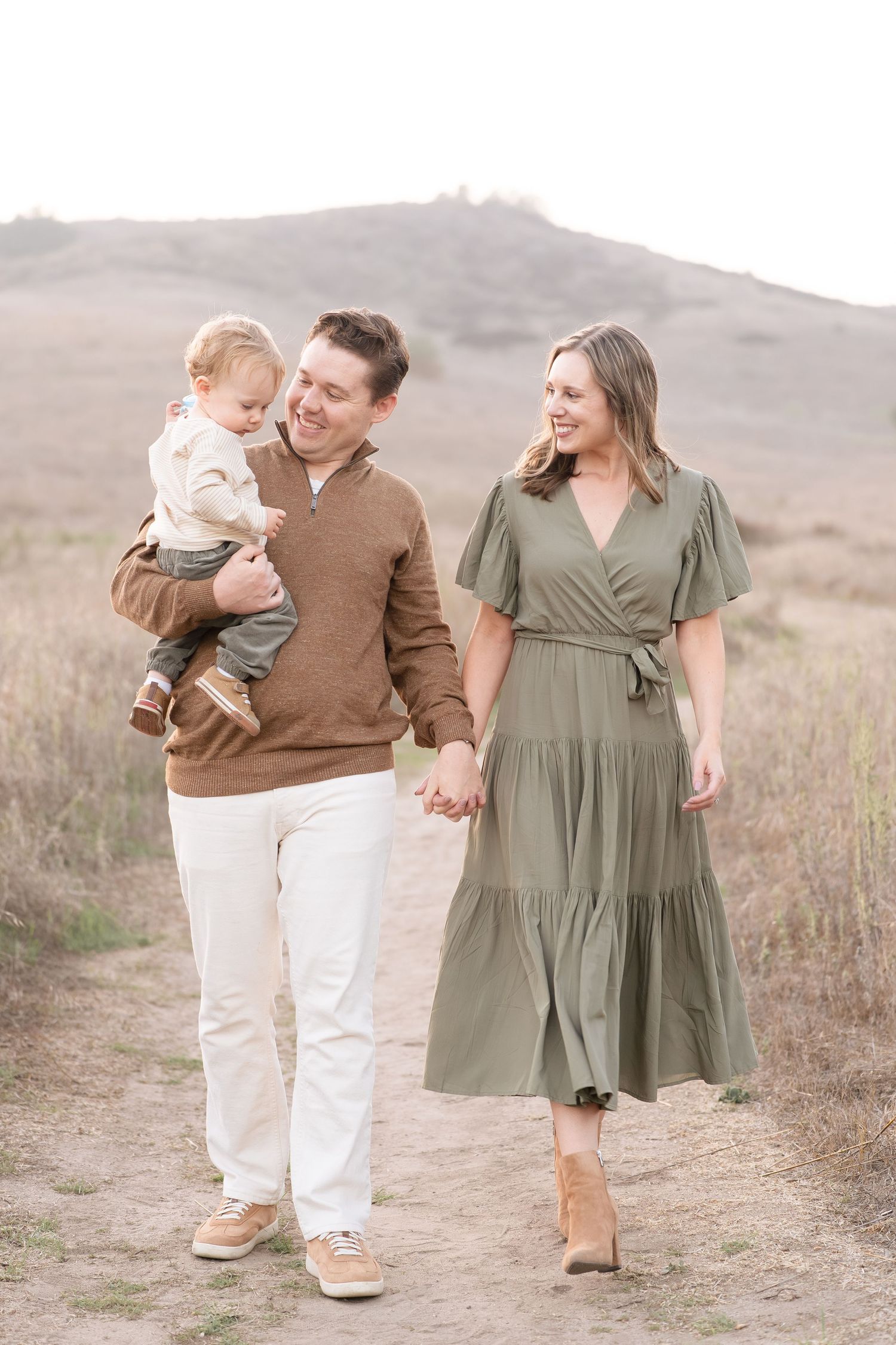 Smiling couple walks hand in hand on a dirt trail, father carrying their young toddler in his arms.