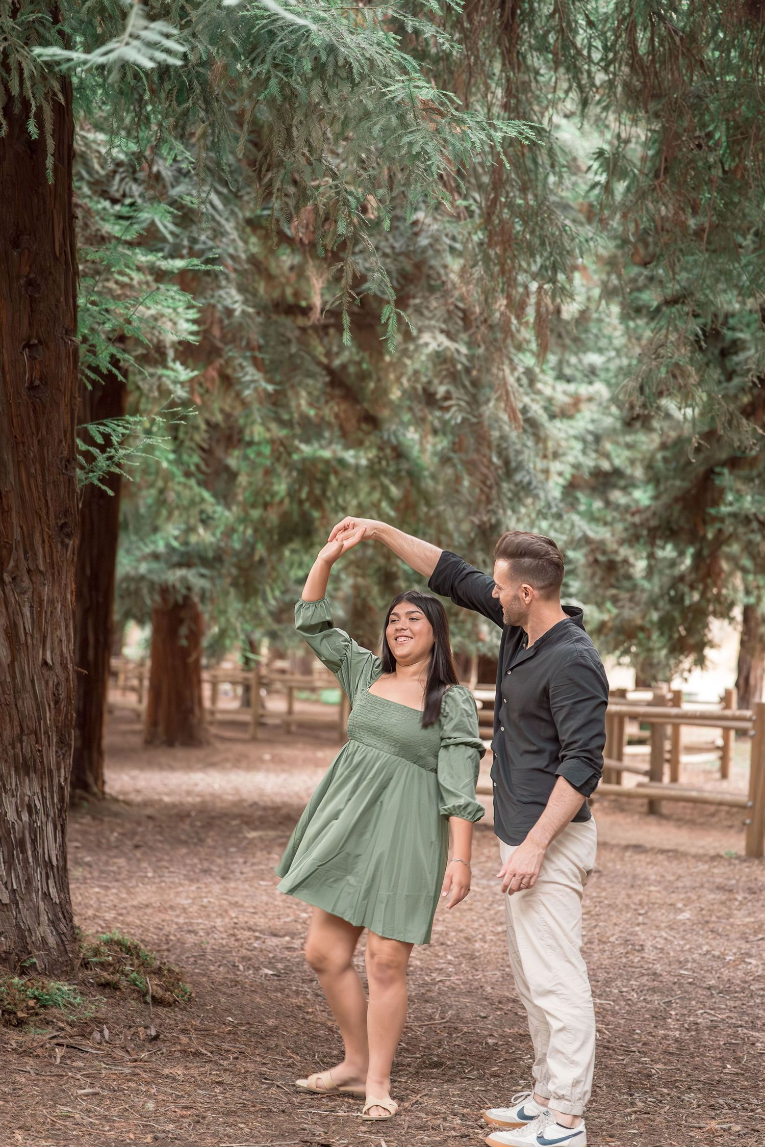 Joyful couple dancing together under towering redwood trees, man spinning woman in green dress.