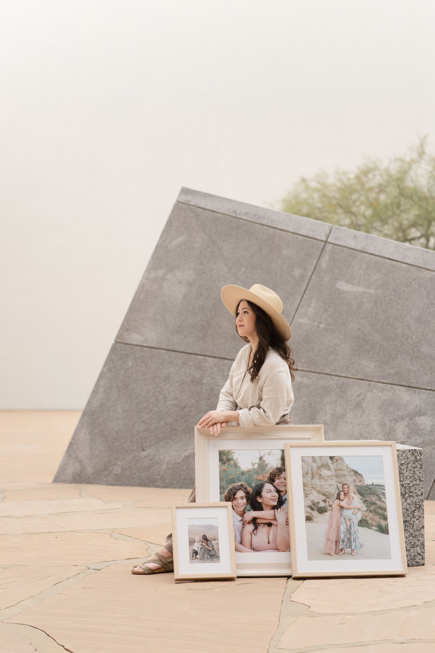 Woman in straw hat sits beside framed family portrait photos against modern stone architecture outdoors.