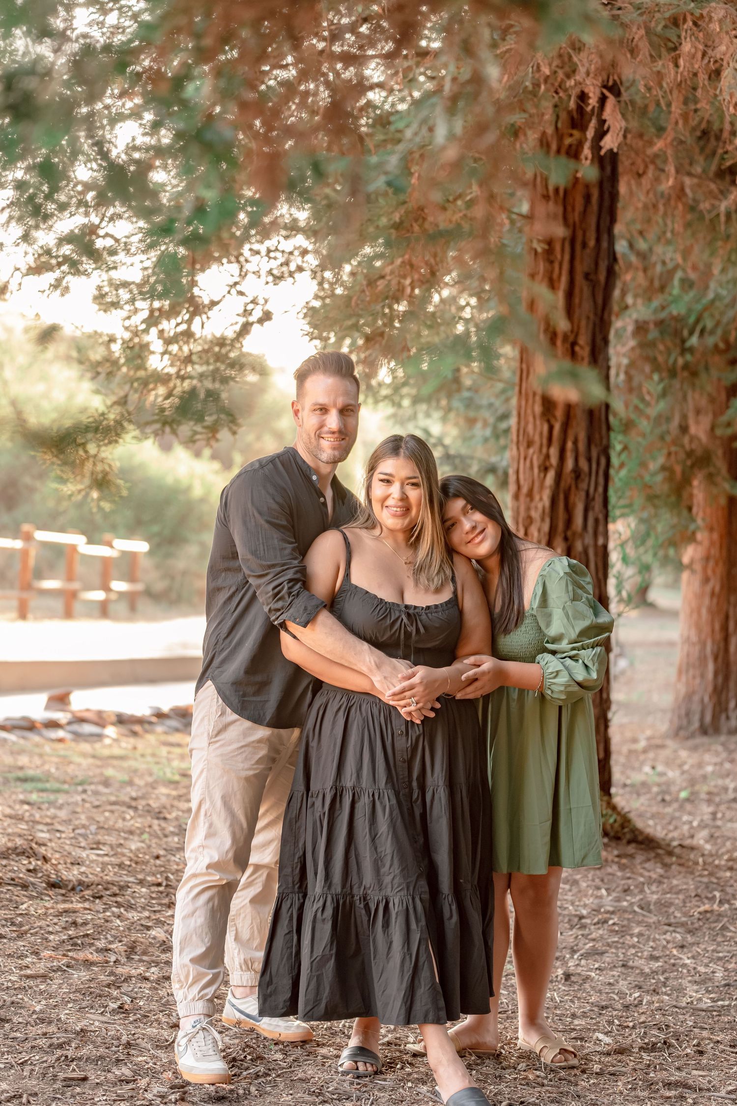 Happy family of three posing together under tall redwood trees in warm golden afternoon light.