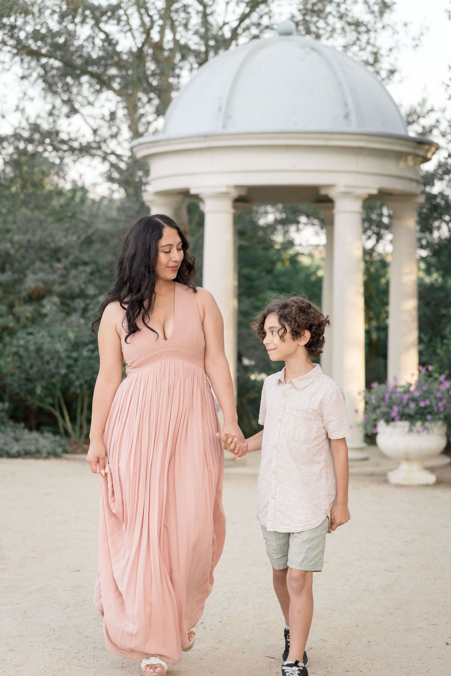 A mother in a blush maxi dress and her curly-haired son walk hand-in-hand near a garden rotunda.