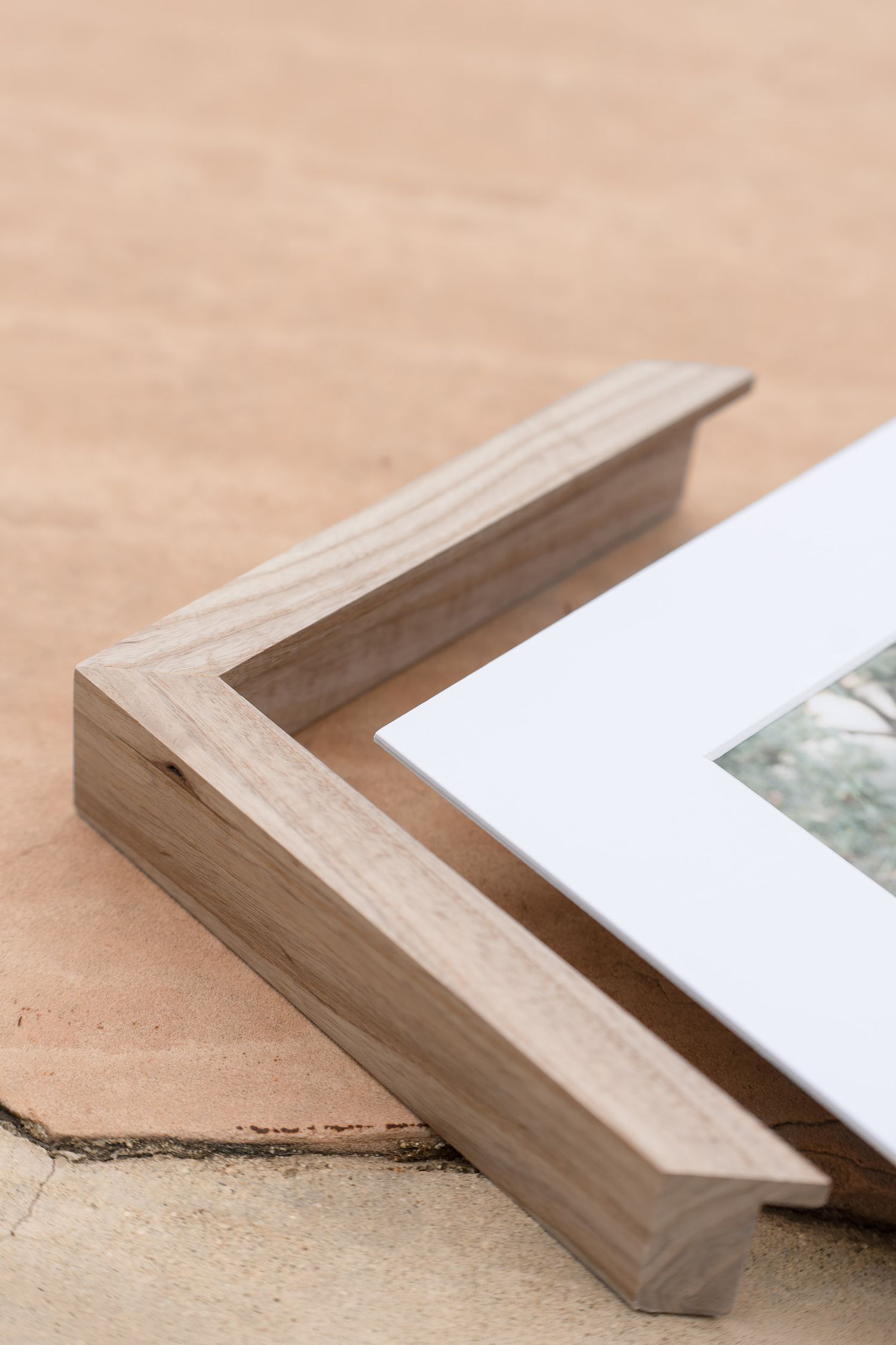 Close-up of a wooden picture frame corner with white mat board on a sandy surface.