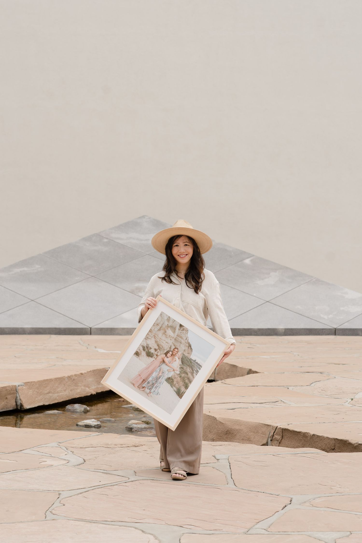 Woman in beige outfit and hat holding a framed photo outdoors on stone pavement near a geometric structure.