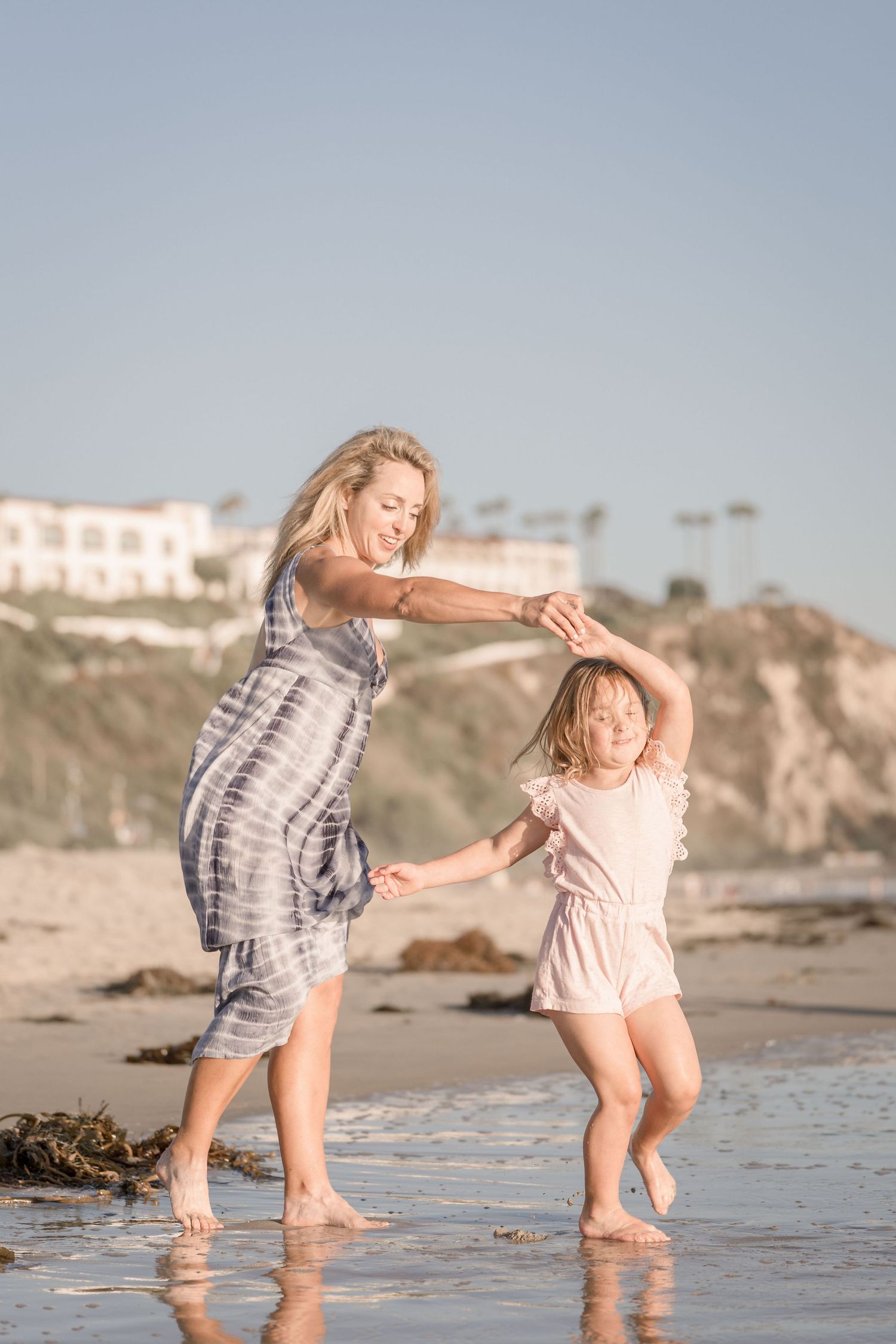 Mother spinning young daughter on wet shoreline beach at sunset in playful moment.
