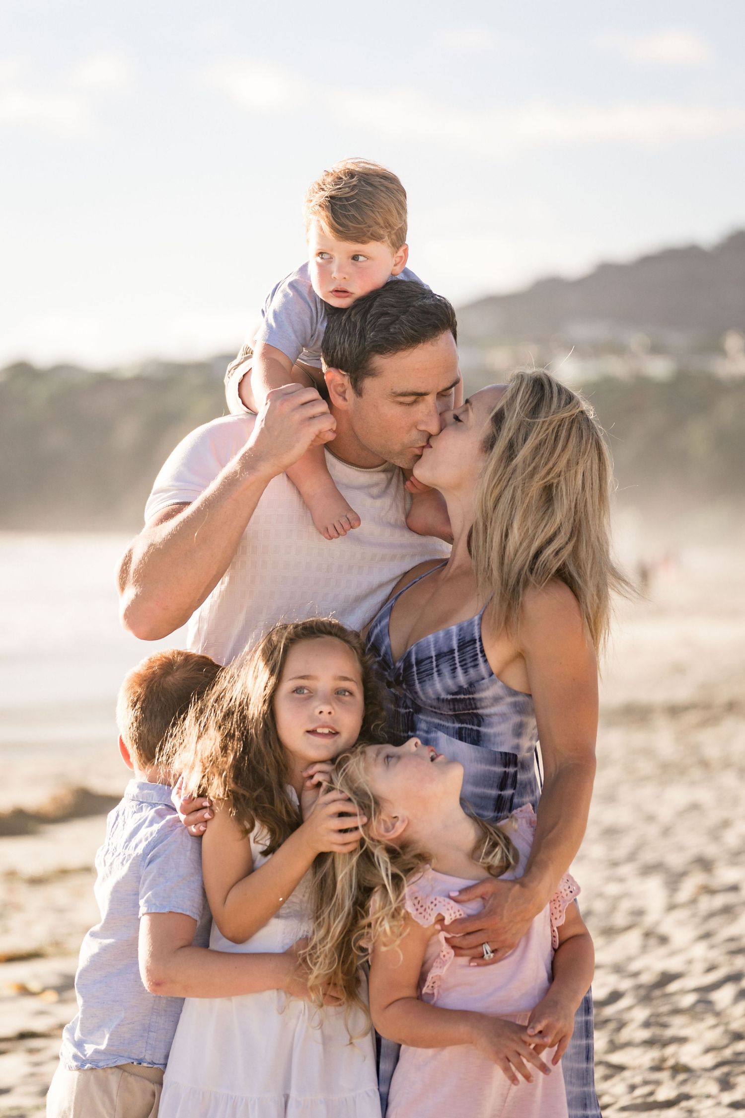 Parents share a kiss on the beach while surrounded by their four young children at sunset.