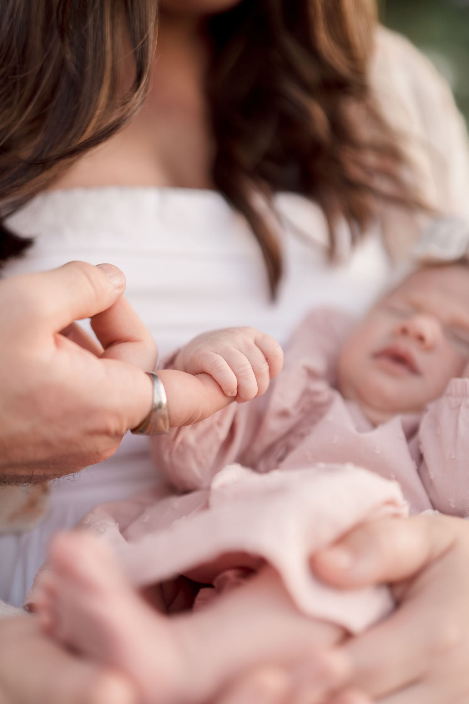 A newborn baby in pink gently grasps a parent's finger, capturing a tender family bonding moment.