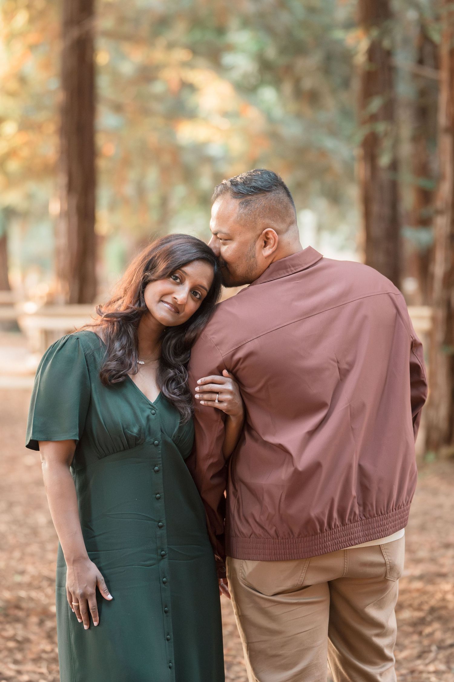 A couple shares a tender moment in a redwood forest, with the man kissing his smiling partner on the cheek.