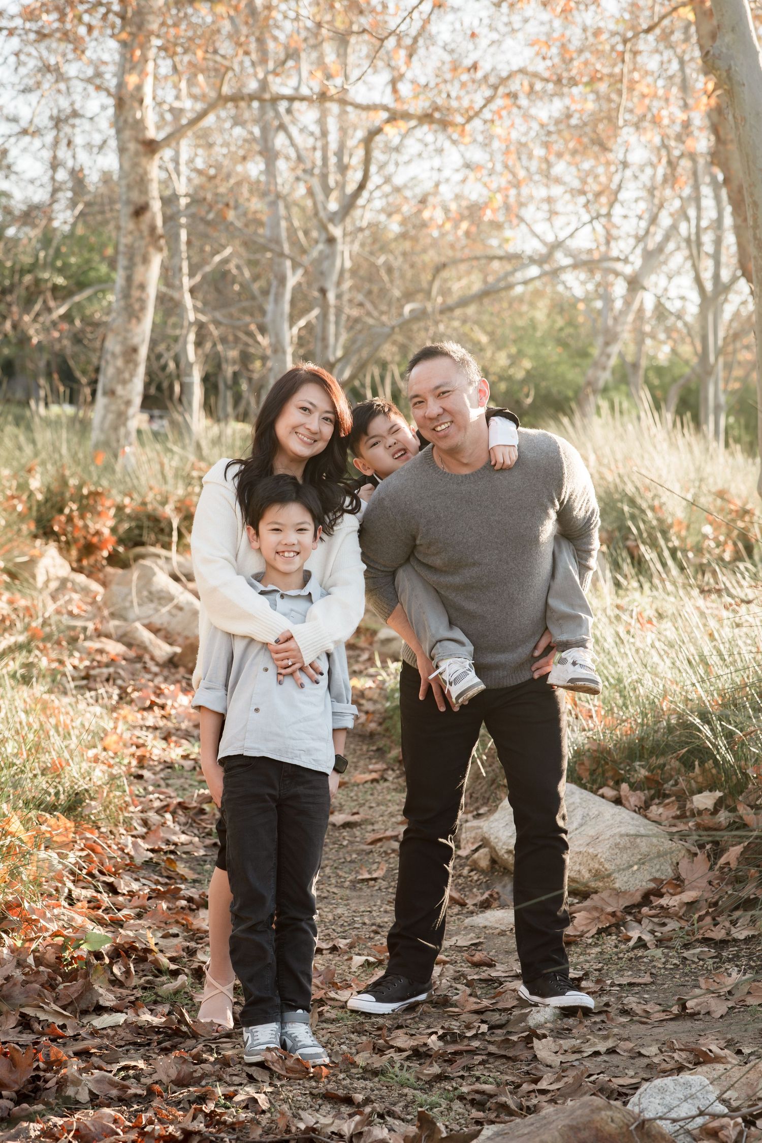 Asian family of four smiling outdoors in autumn park with fallen leaves and bare trees.