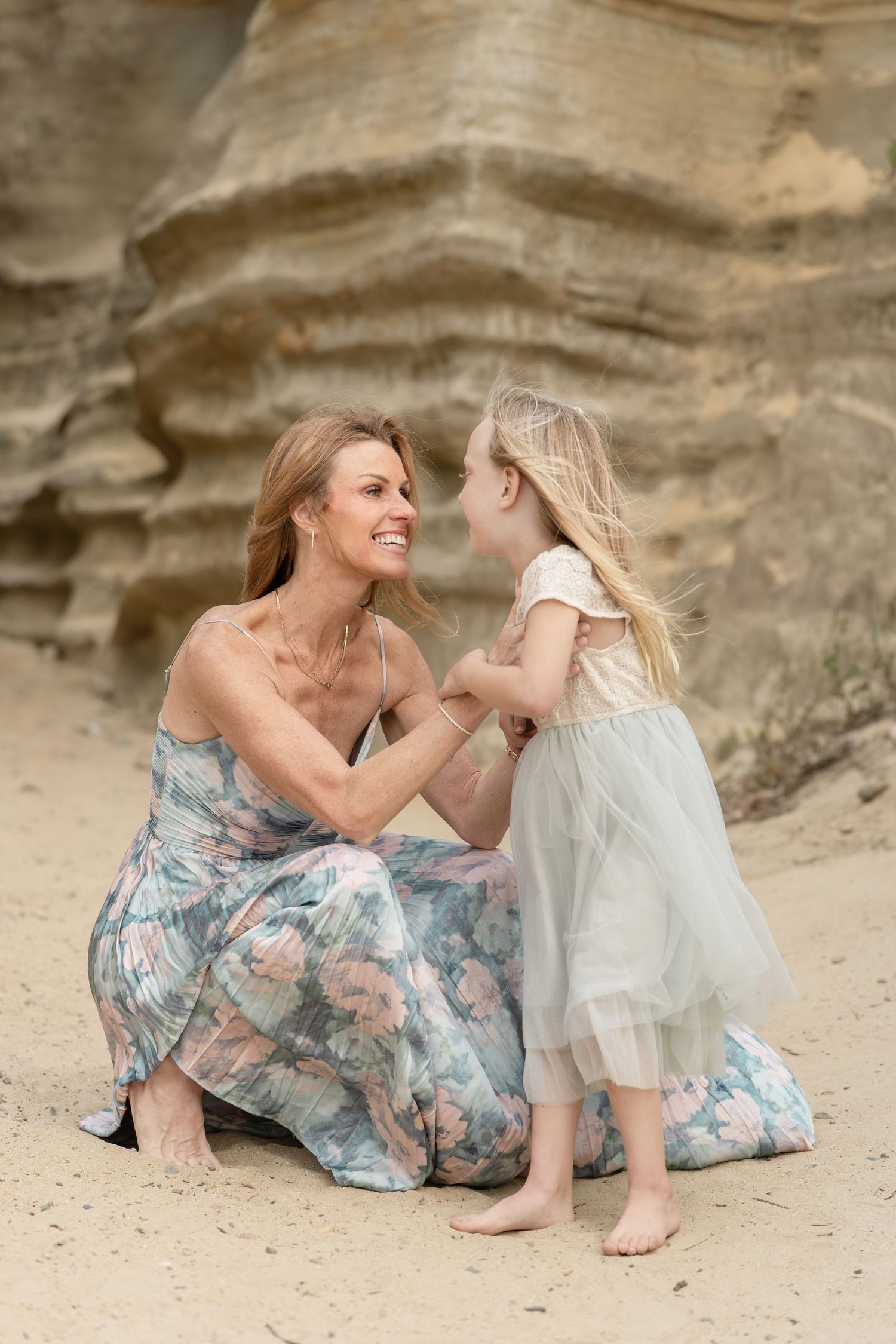 A smiling woman in a floral dress sits on sand with a young blonde girl in a tulle dress near rocky cliffs.