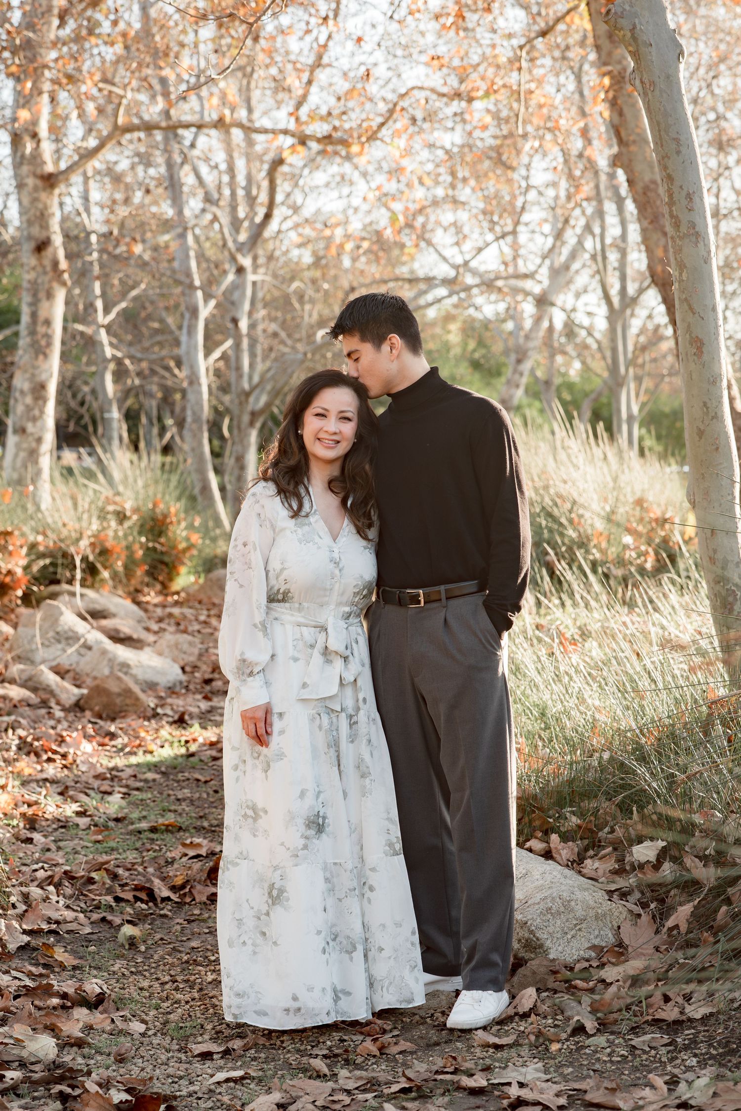 Couple embracing in autumn park, woman in floral dress, man in black sweater, surrounded by fall foliage.