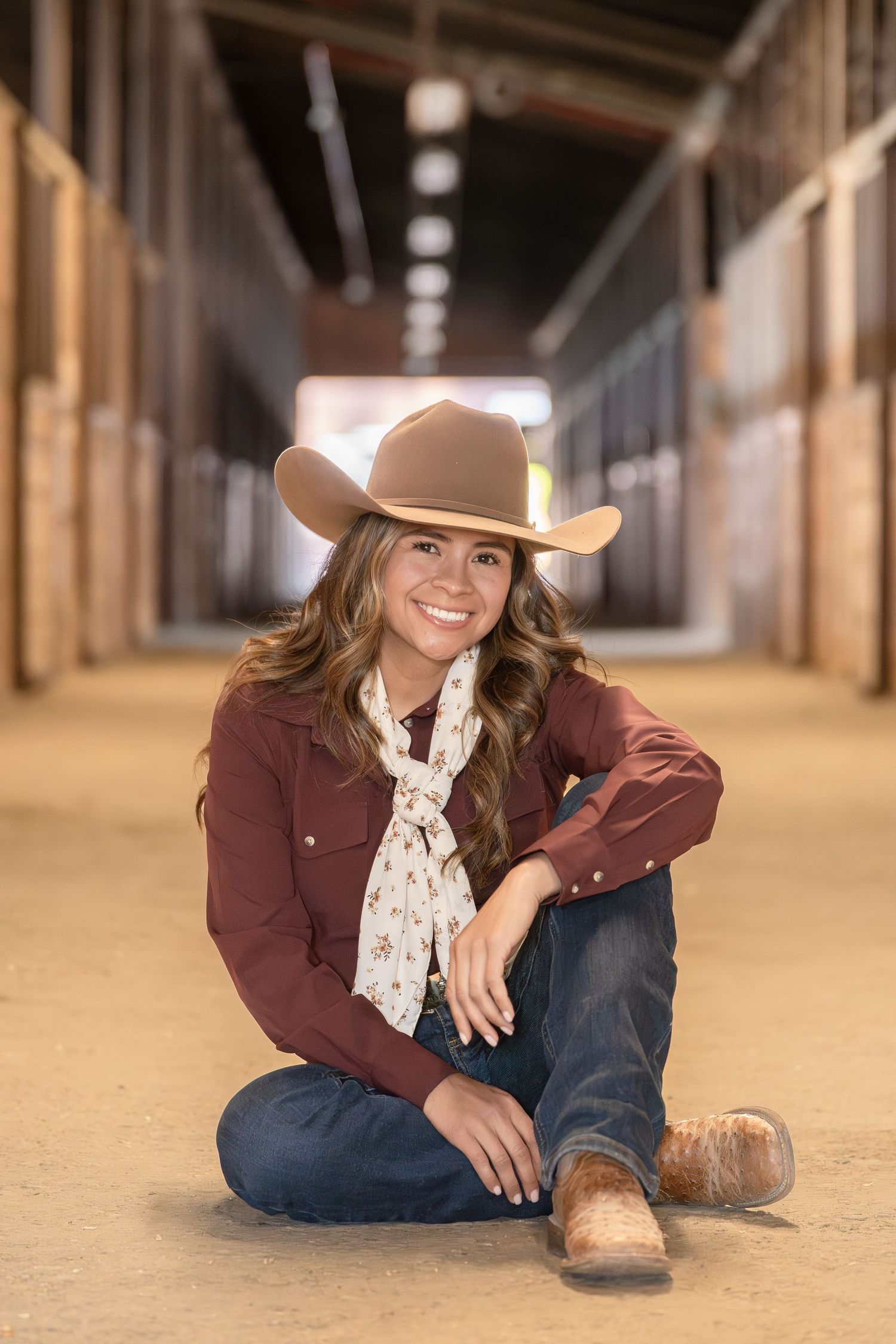 Smiling young woman in cowboy hat, burgundy shirt, jeans, and boots sitting cross-legged in a horse stable aisle.