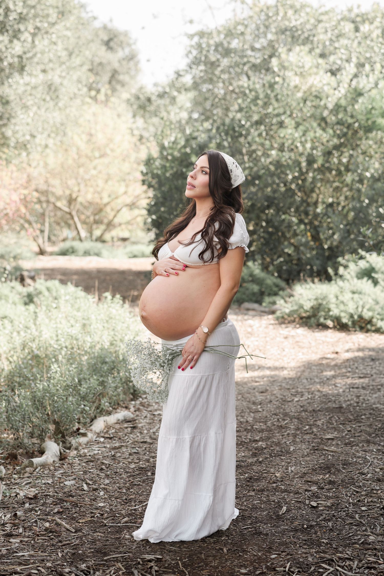 Pregnant woman in white dress cradling baby bump outdoors in a lush garden setting during a maternity photoshoot.