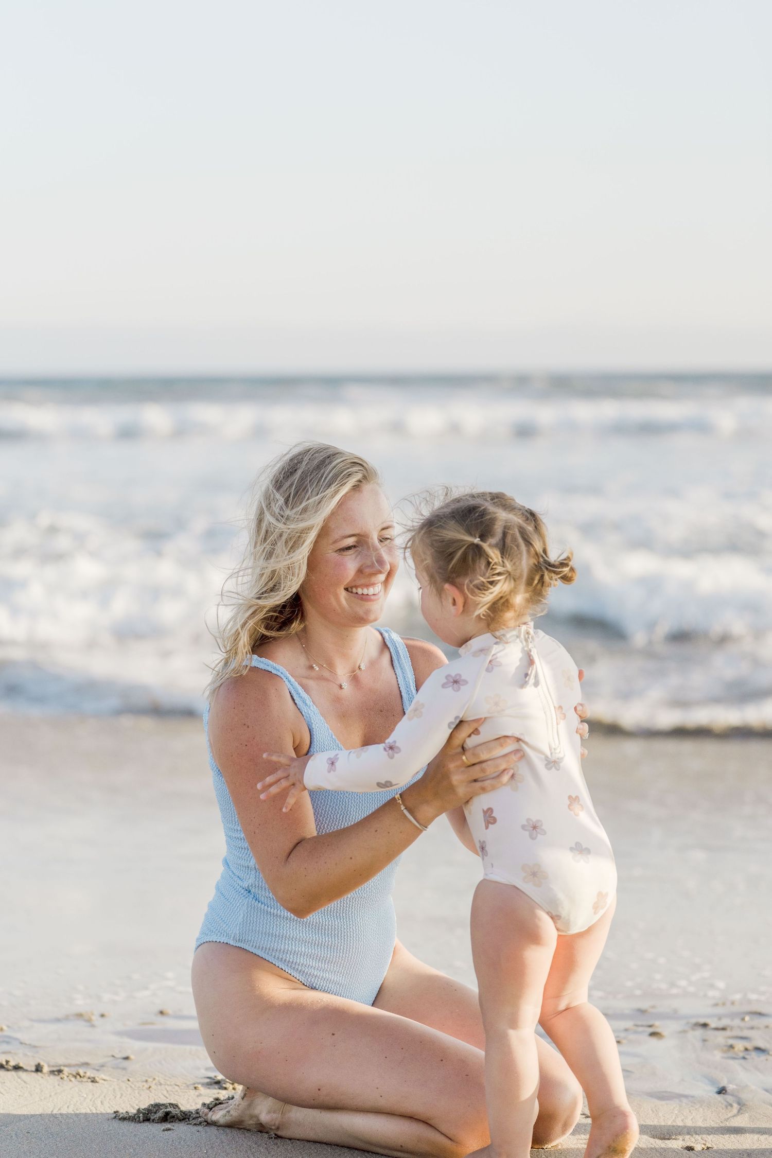 Smiling blonde mother in blue swimsuit holds toddler daughter at ocean beach shoreline.