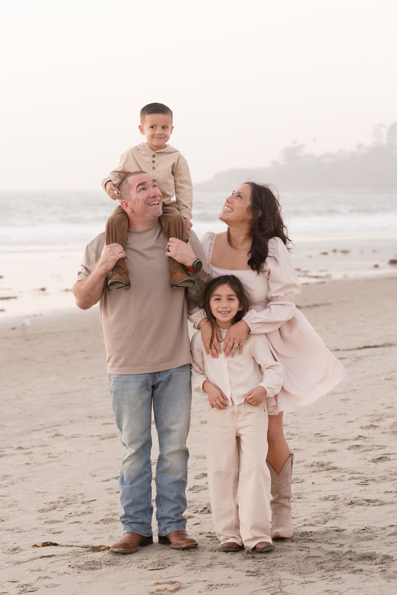 Happy family of four poses on a sandy beach at dusk, father with son on shoulders, mother hugging daughter.