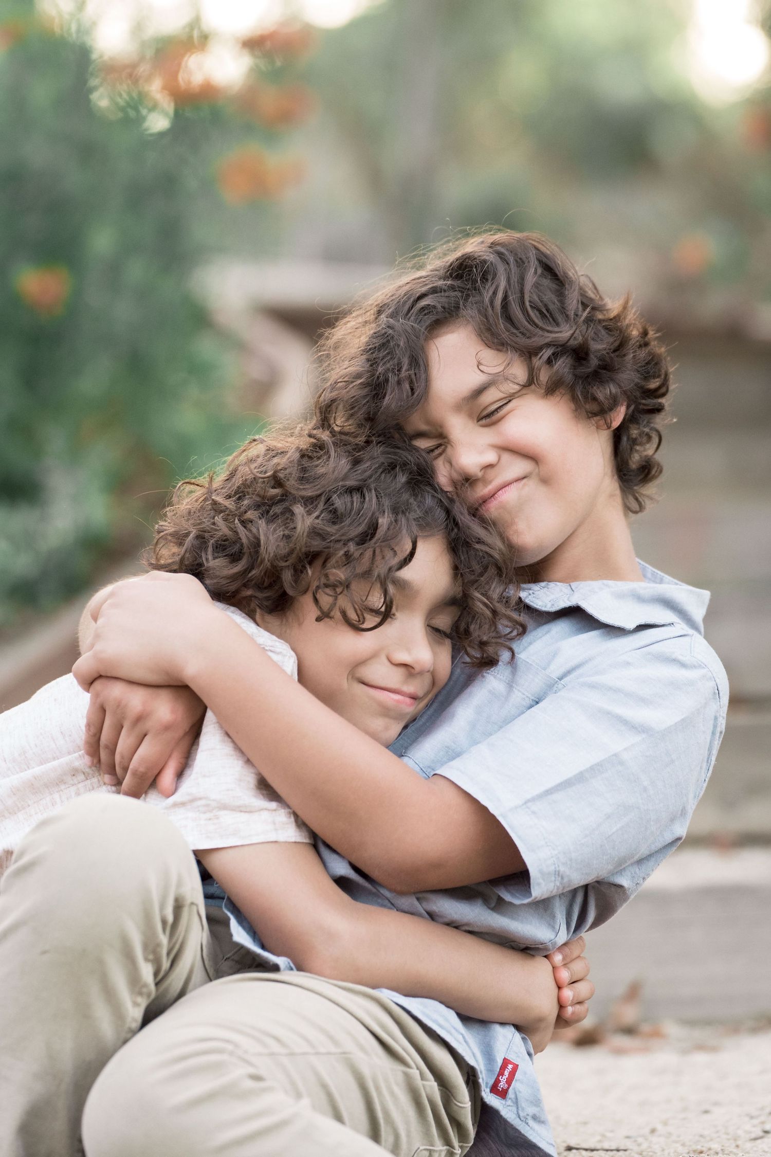 Two curly-haired brothers sharing a warm hug while sitting outdoors in a sunlit garden setting.