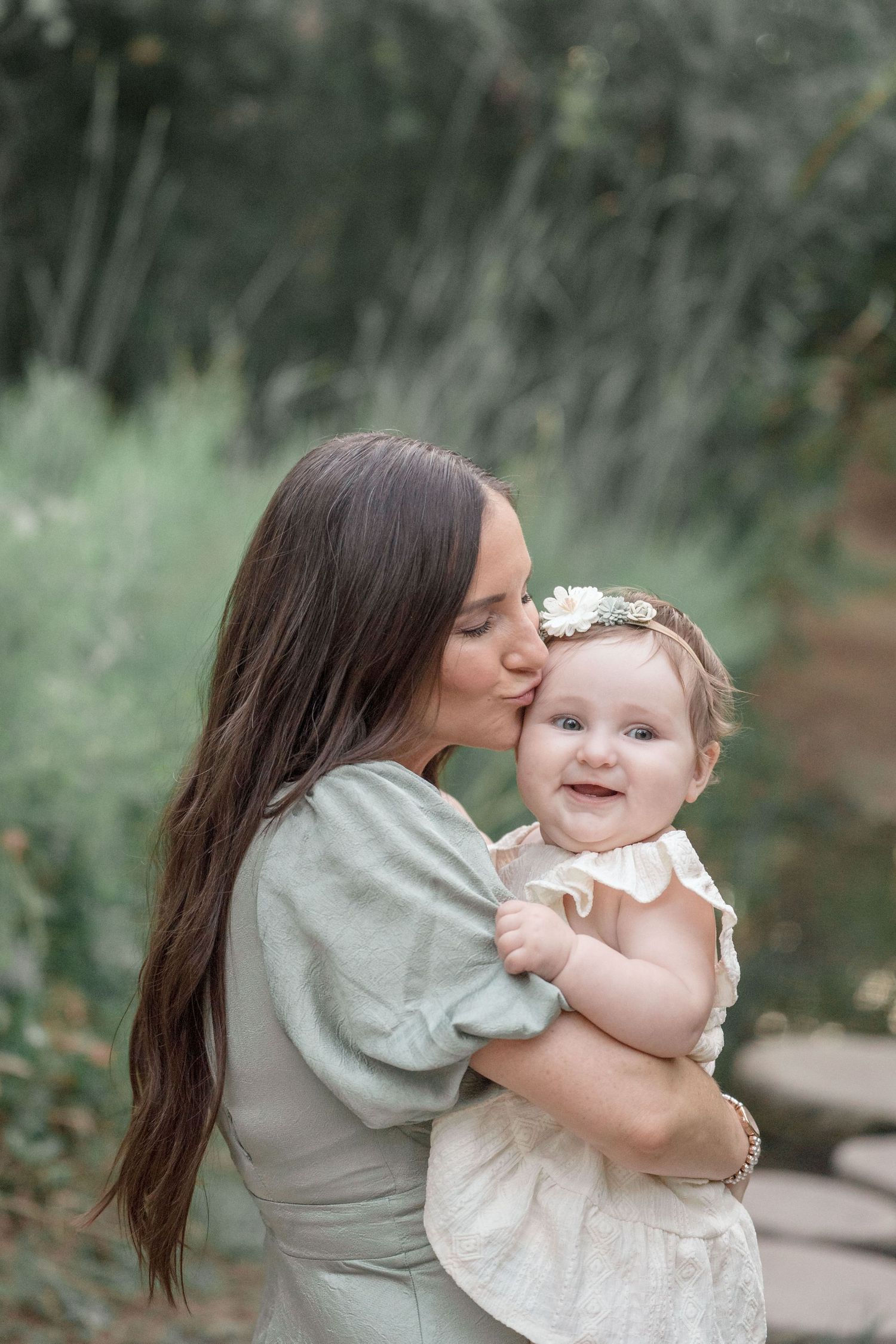 Mother tenderly kissing smiling baby girl wearing a floral headband in a lush outdoor setting.