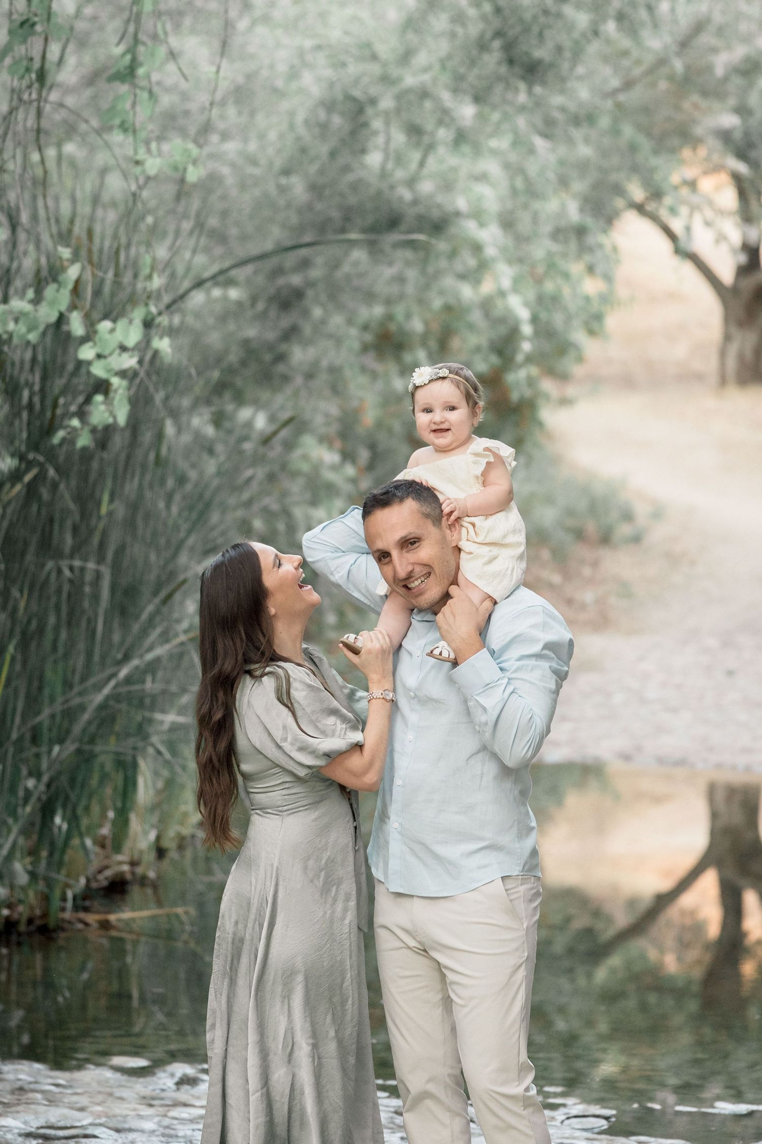 Happy family with father holding baby on shoulders beside a peaceful pond surrounded by greenery.