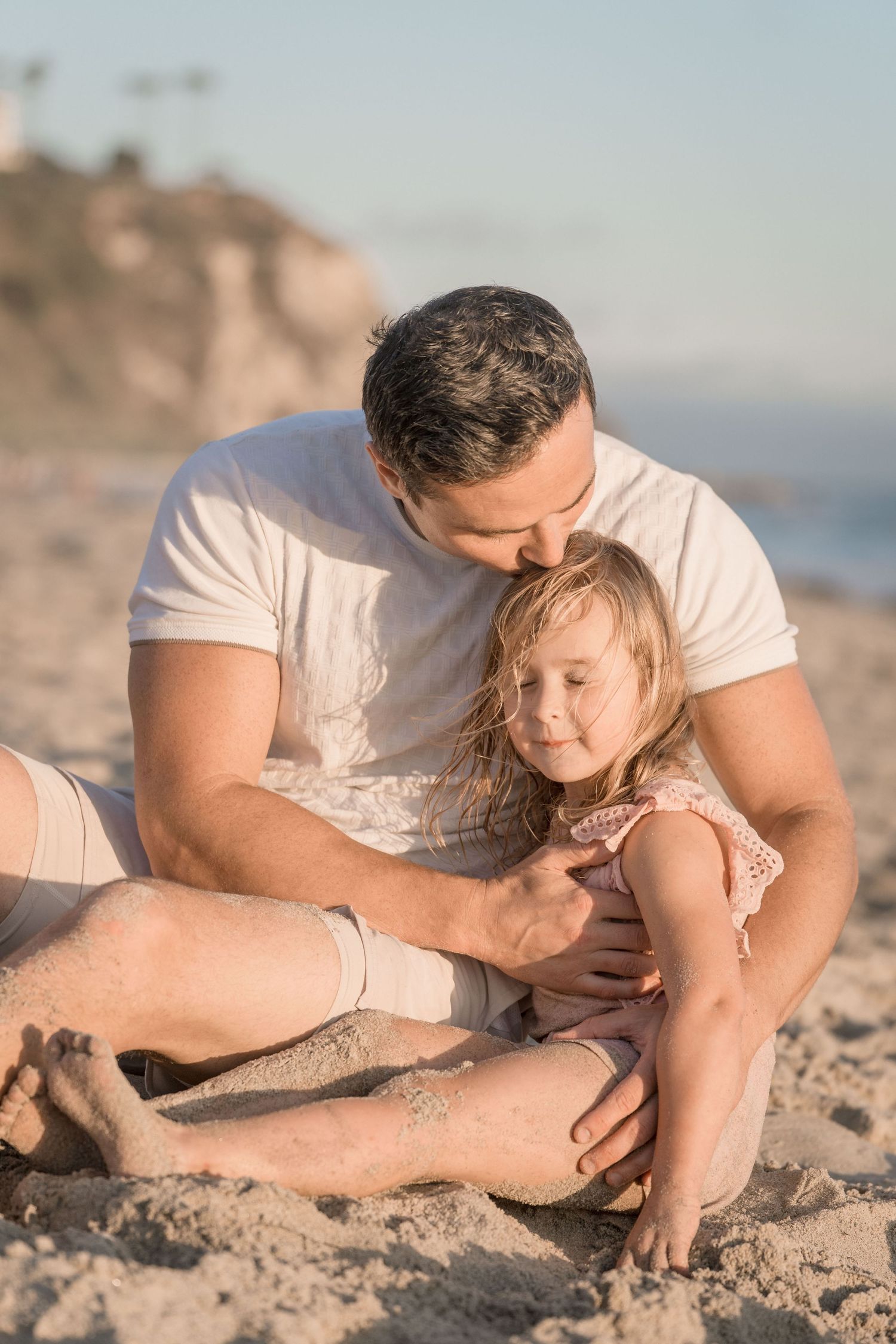 Father sitting on sandy beach cradling young blonde daughter in warm sunset light.