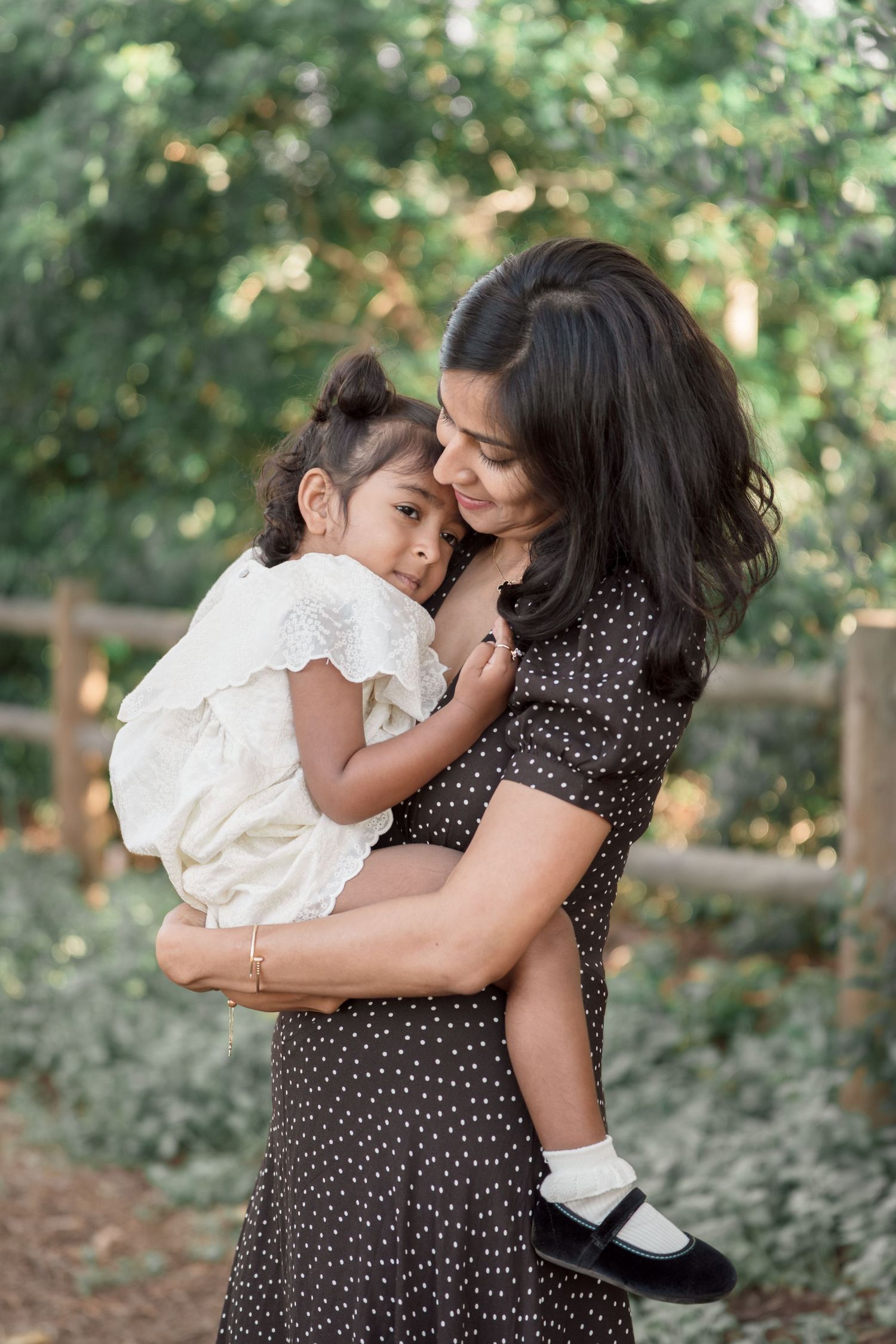 A mother in a polka dot dress tenderly holds her young daughter outdoors near a wooden fence with lush greenery.