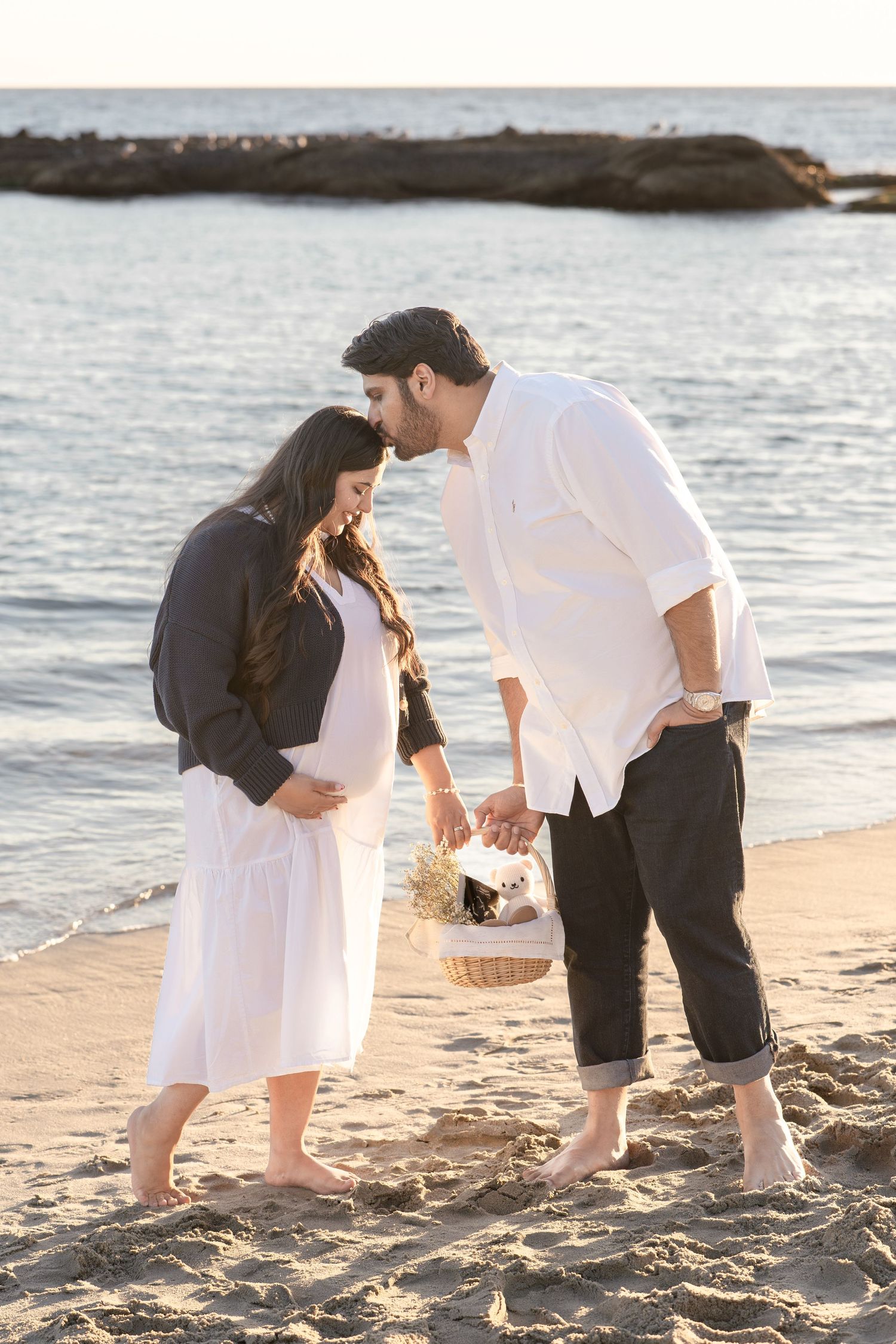 Expecting couple standing barefoot on sandy beach at sunset, man kissing pregnant womans forehead.