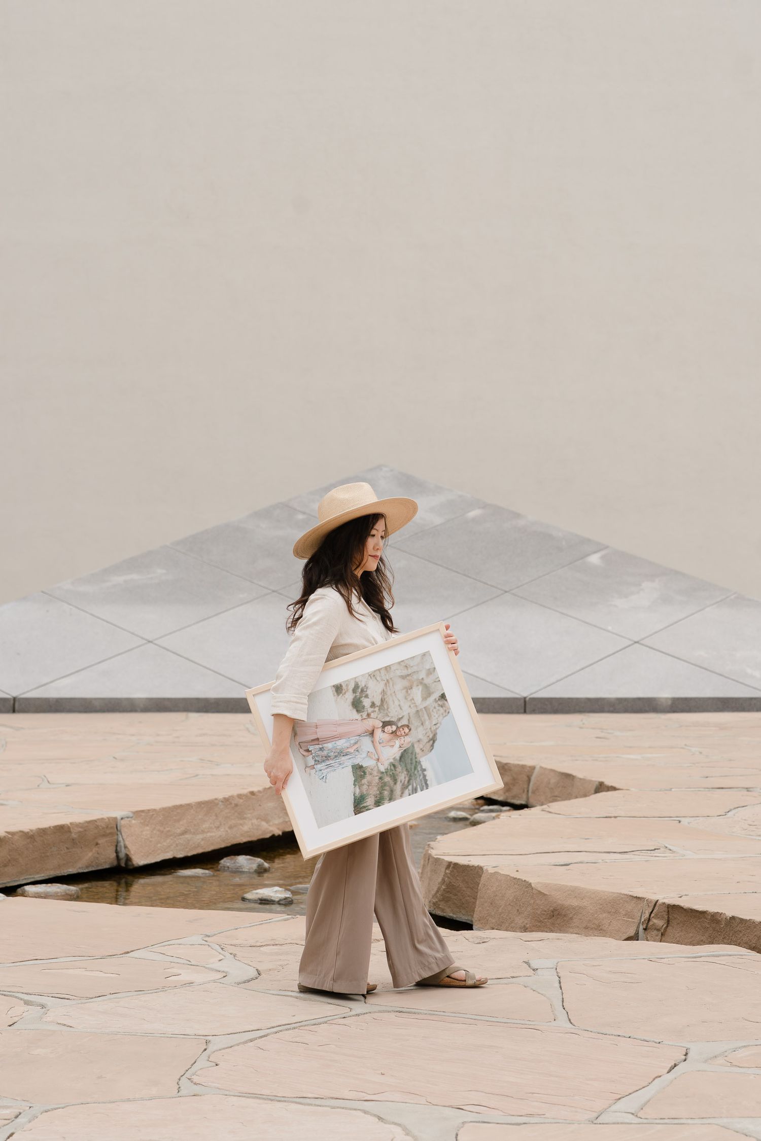 Woman in straw hat carrying framed artwork across a stone plaza with geometric water feature.