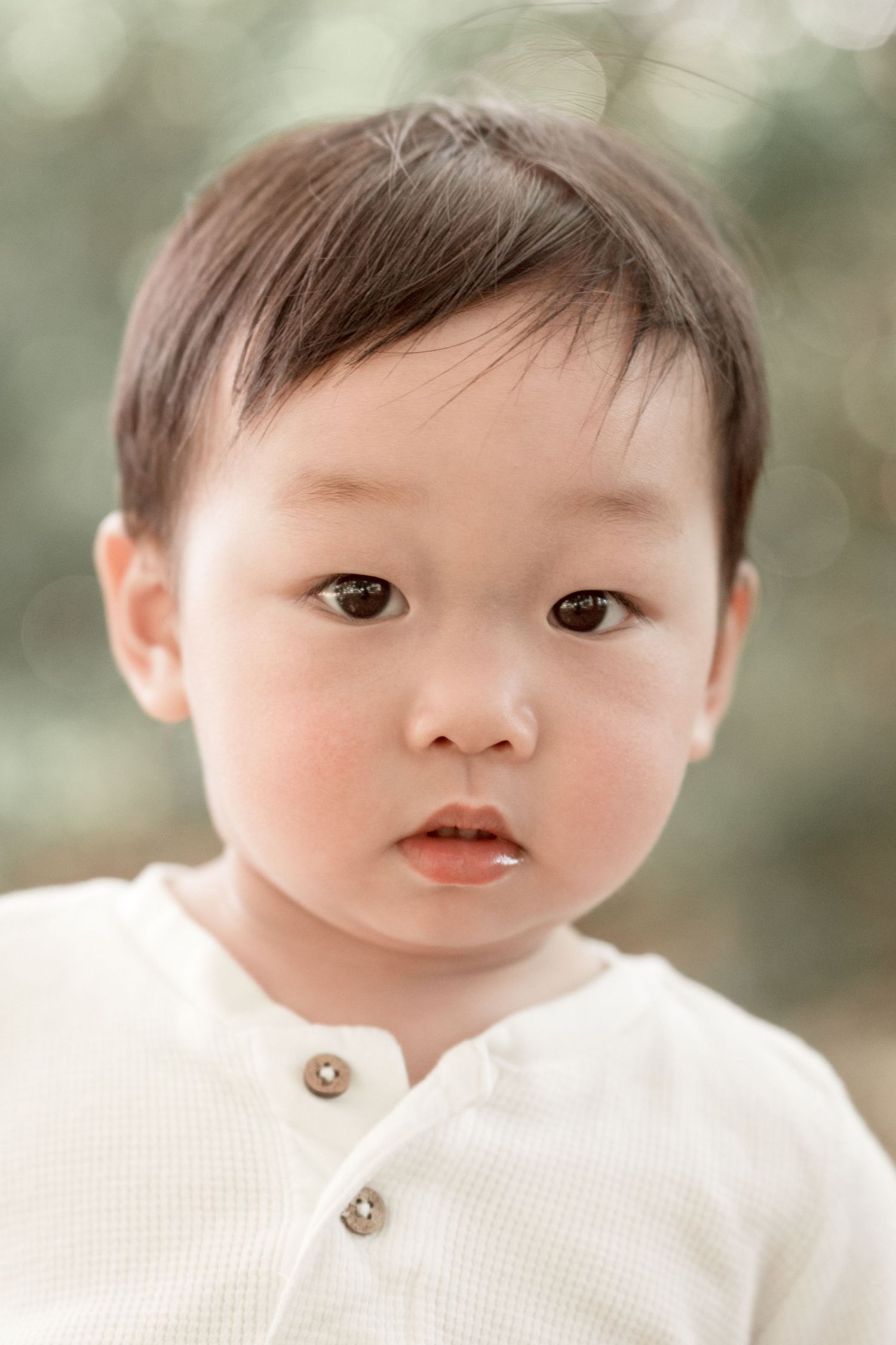Adorable Asian toddler with dark eyes wearing a white henley shirt, photographed outdoors with soft bokeh background.