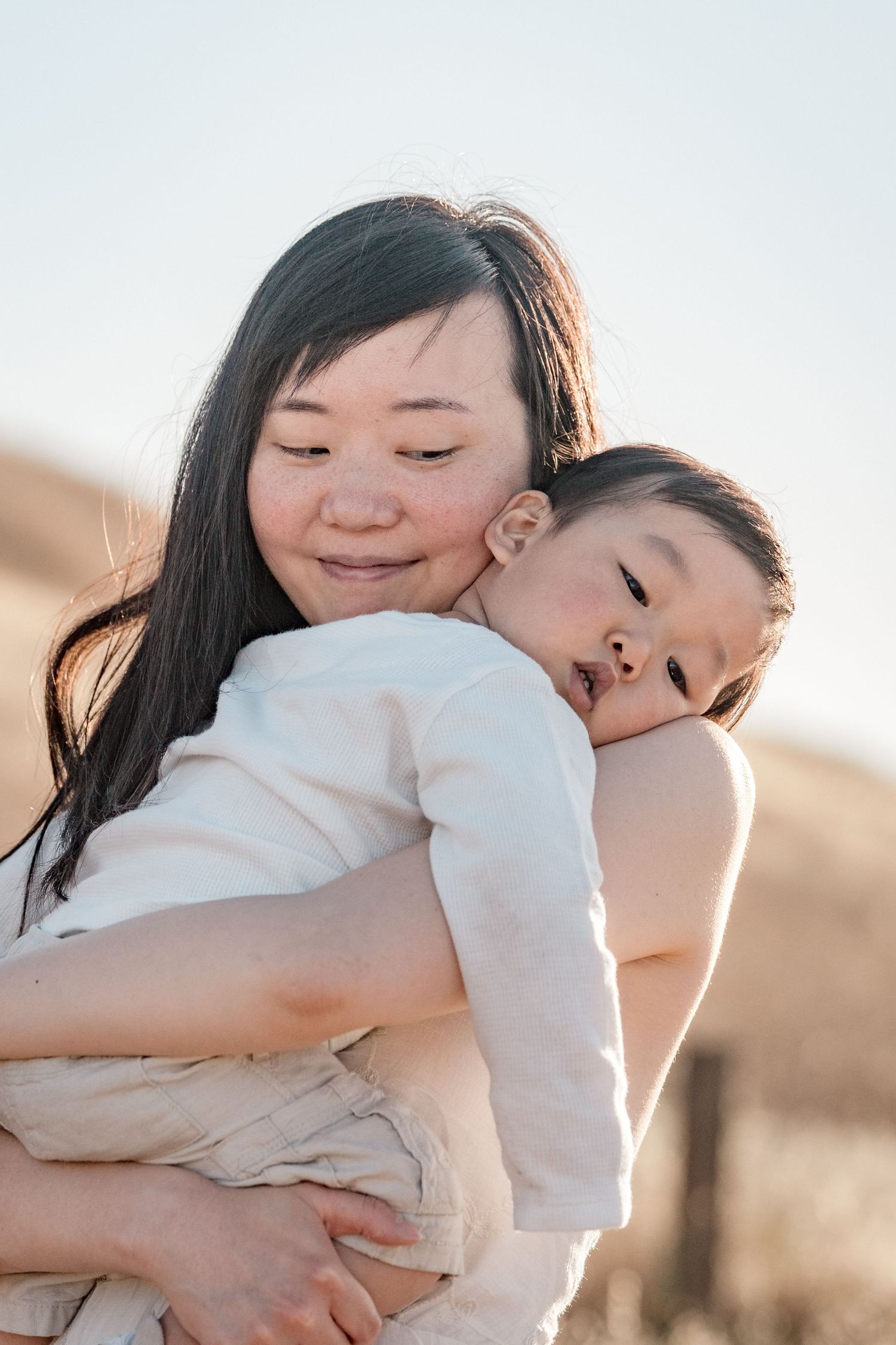 A smiling Asian mother tenderly holds her sleepy toddler outdoors in warm golden light.