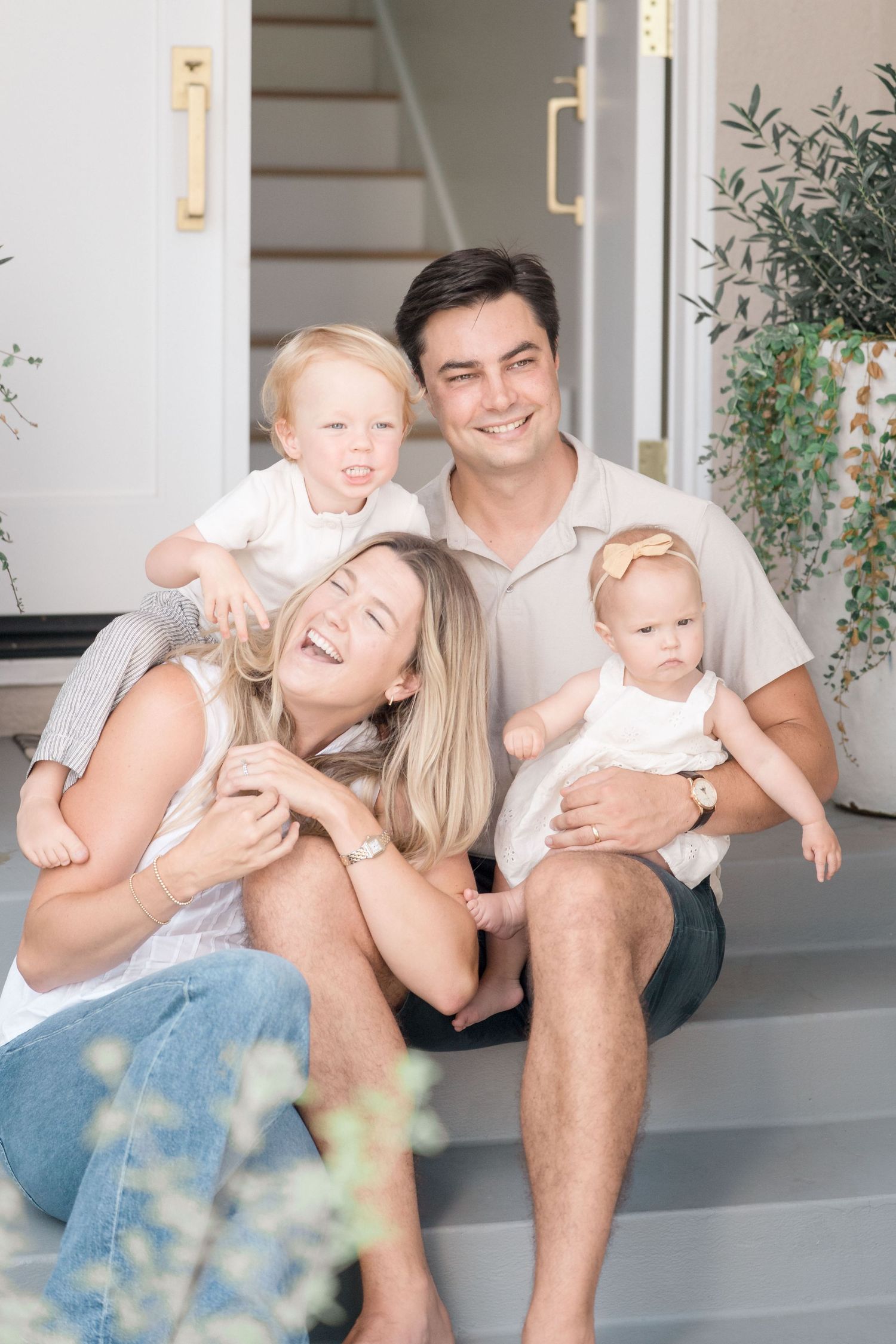 A cheerful family of four sits on front porch steps, parents laughing with their toddler and baby.
