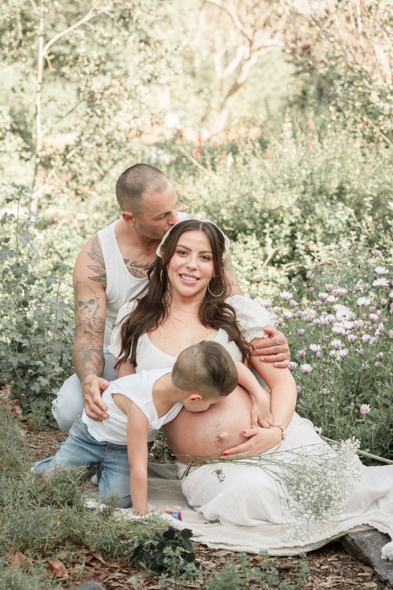 A pregnant woman sits outdoors with her partner and young child in a flower-filled garden during a maternity photoshoot.