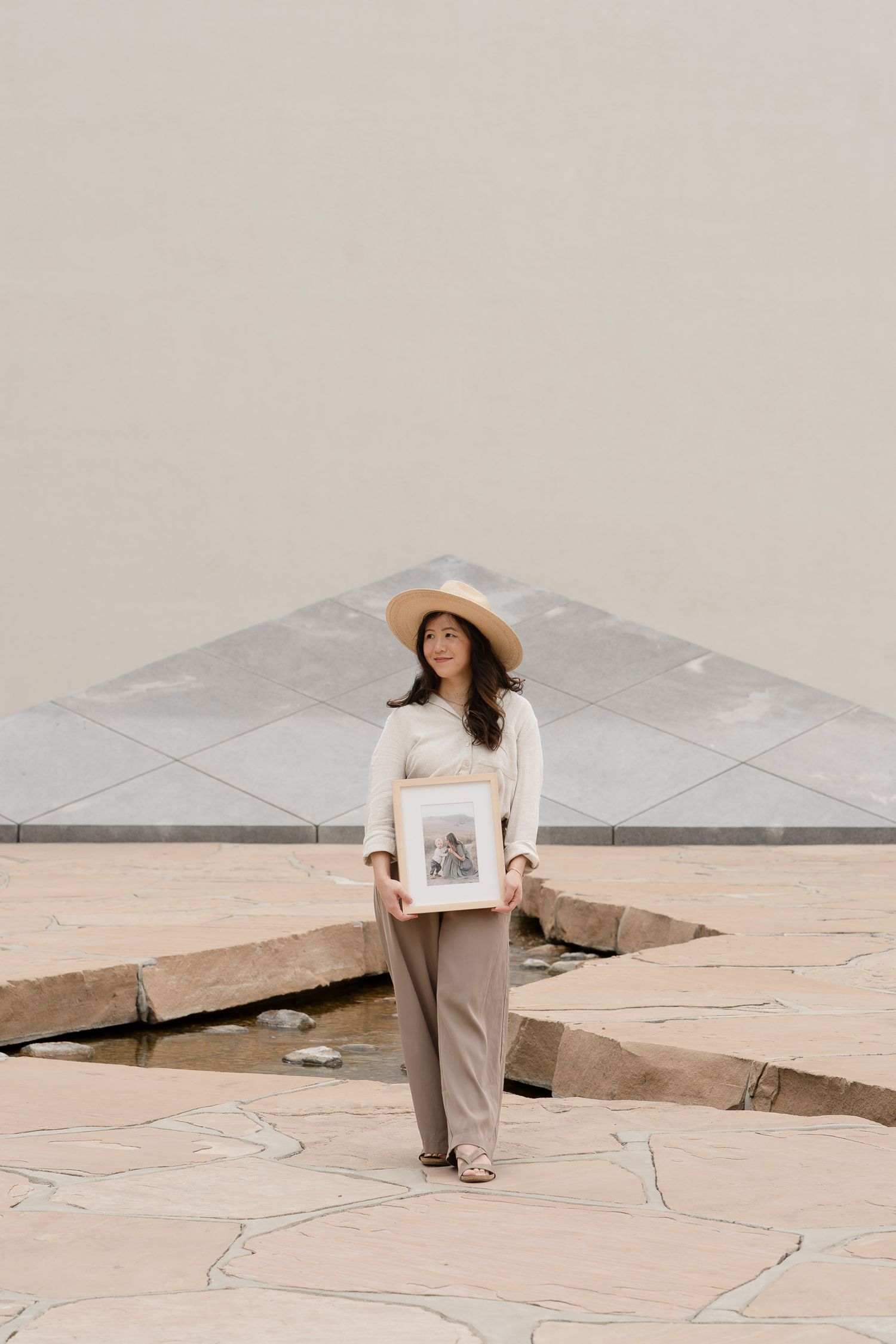Woman in beige outfit and hat holds framed artwork outdoors near a geometric water feature.